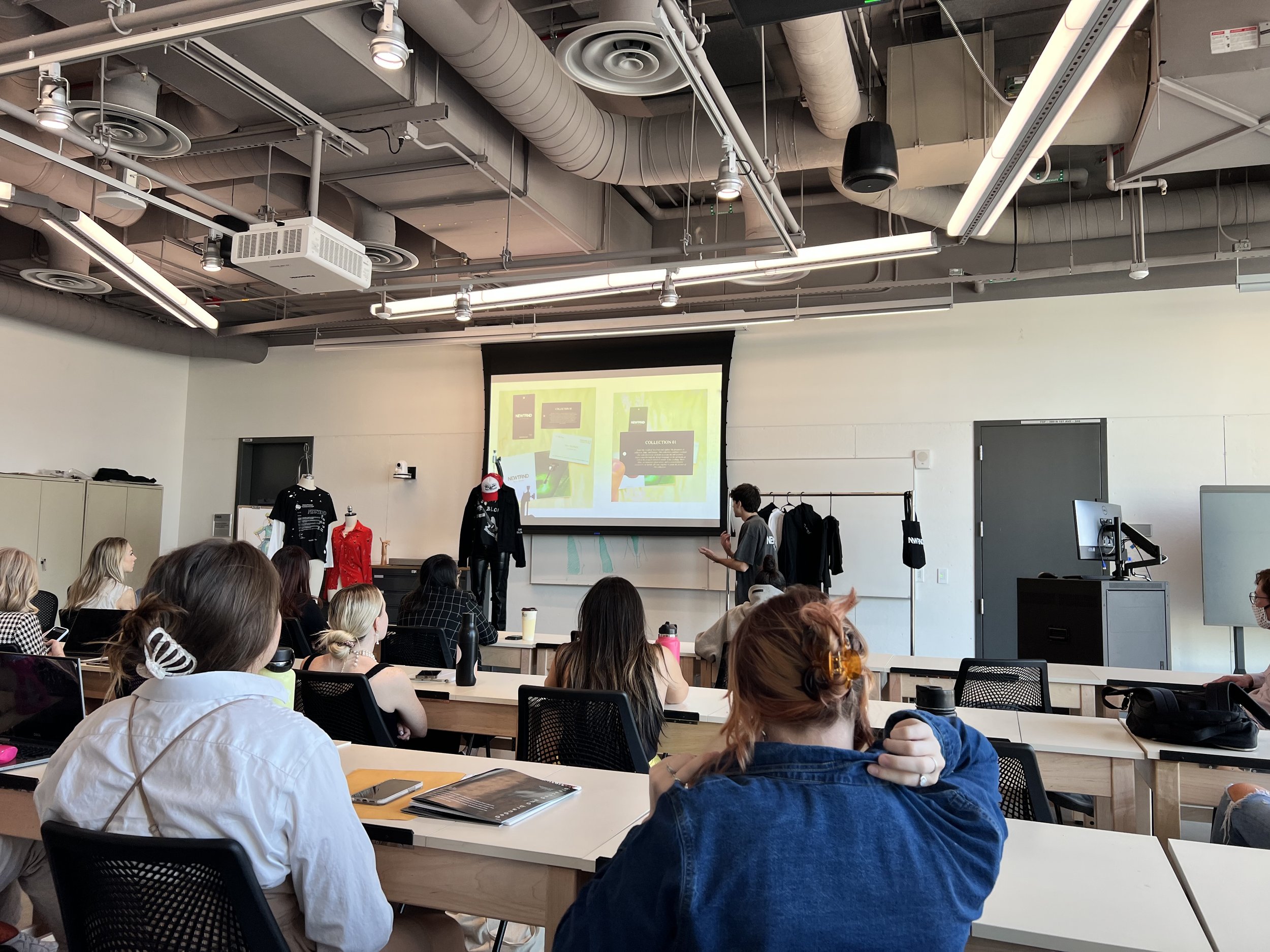 A classroom or seminar room filled with students listening to a speaker at the front, with a projection screen displaying a presentation, and clothing mannequins with garments on display.