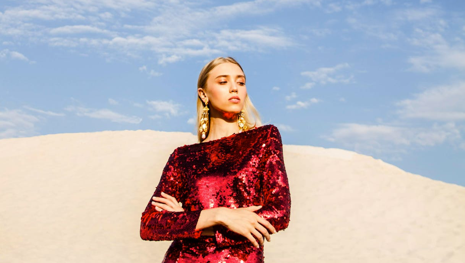A woman in a red sequined dress with gold earrings standing outdoors with a cloudy sky and sandy landscape in the background.