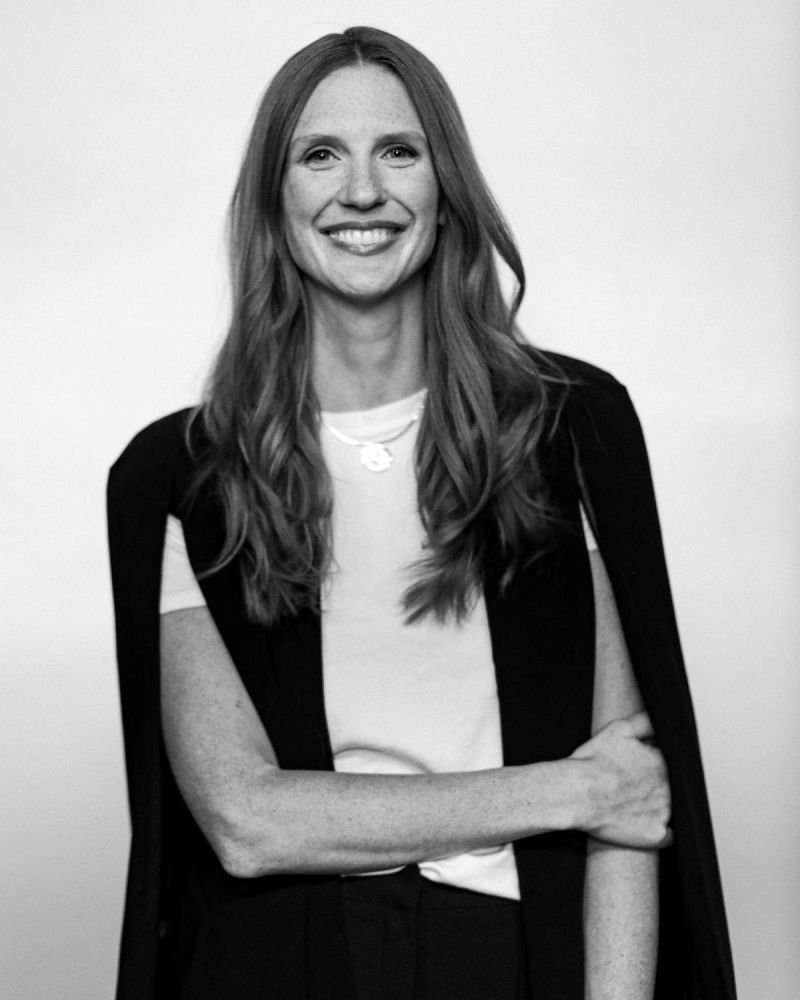 Black and white photo of a smiling woman with long hair, wearing a white top and a dark blazer, standing with her arms crossed against a plain background.