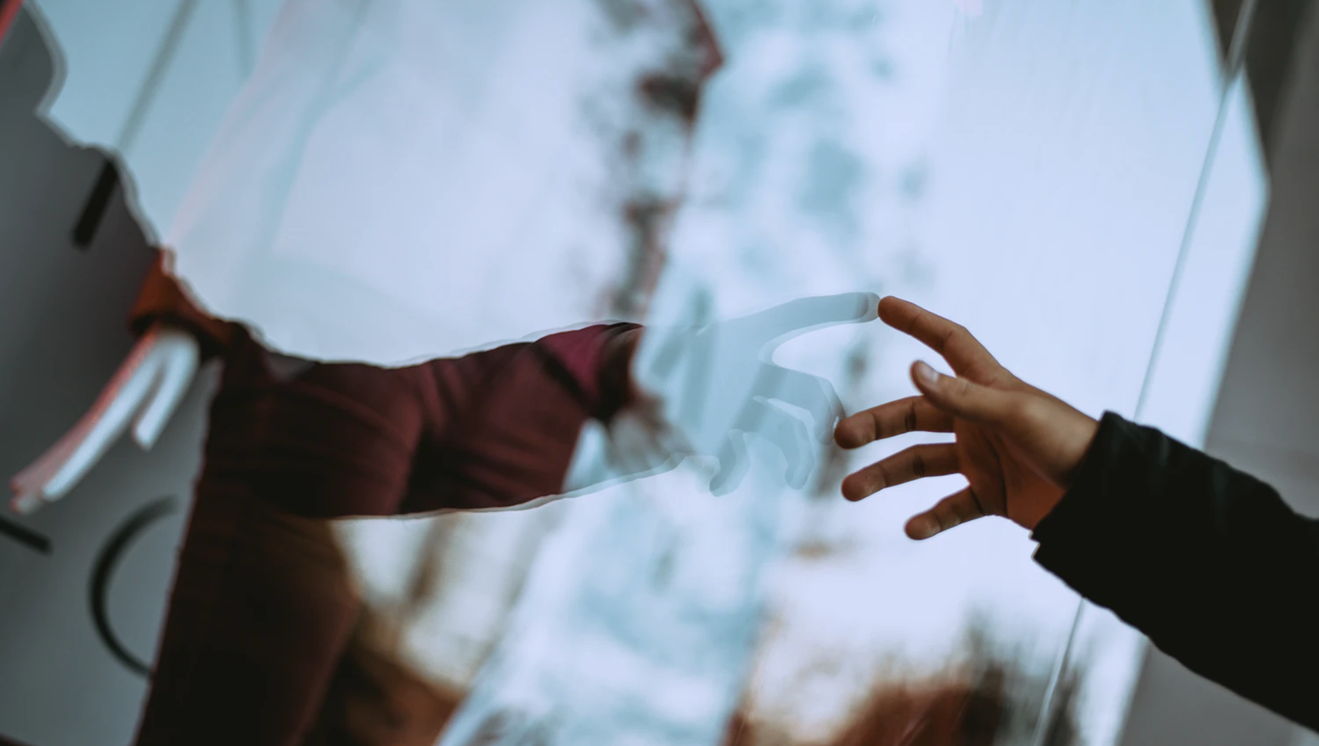 A person touching a reflective glass surface that shows a blurred reflection of their hand and a blue sky with clouds.