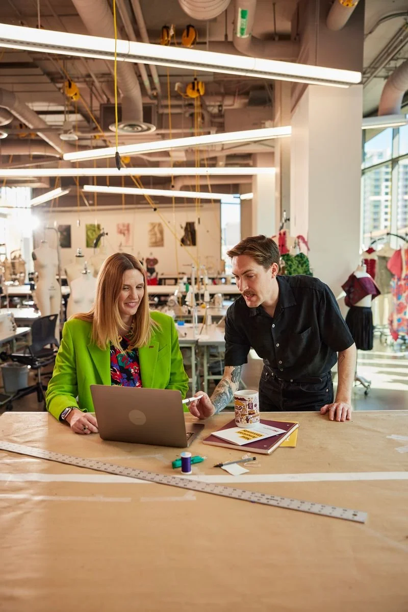 Two people working together at a sewing or fashion design workshop in a creative studio. The woman seated is using a laptop, and the man standing is handing her a piece of fabric or paper. The workspace includes sewing patterns, fabric, and measuring tools, with mannequins and dress designs in the background.