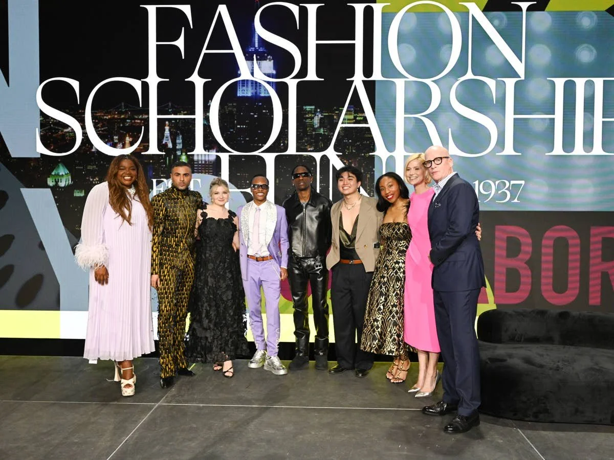 A group of nine diverse people standing on stage in front of a large event banner that reads "Fashion Scholarship". They are dressed in fashionable clothing and are smiling for the photo, with a city skyline backdrop on the banner.