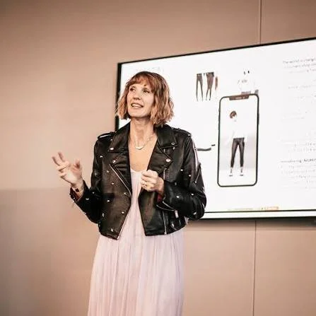 Woman with short red hair wearing a black leather jacket and pink dress standing in front of a large screen.