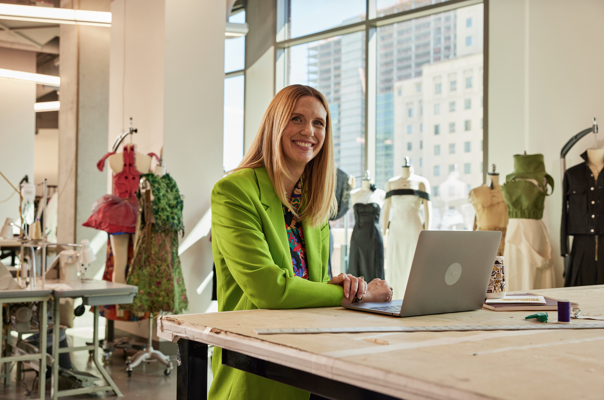 A woman with red hair wearing a bright green blazer sitting at a large wooden table in a modern, well-lit room with large windows and mannequins displaying fashion designs.