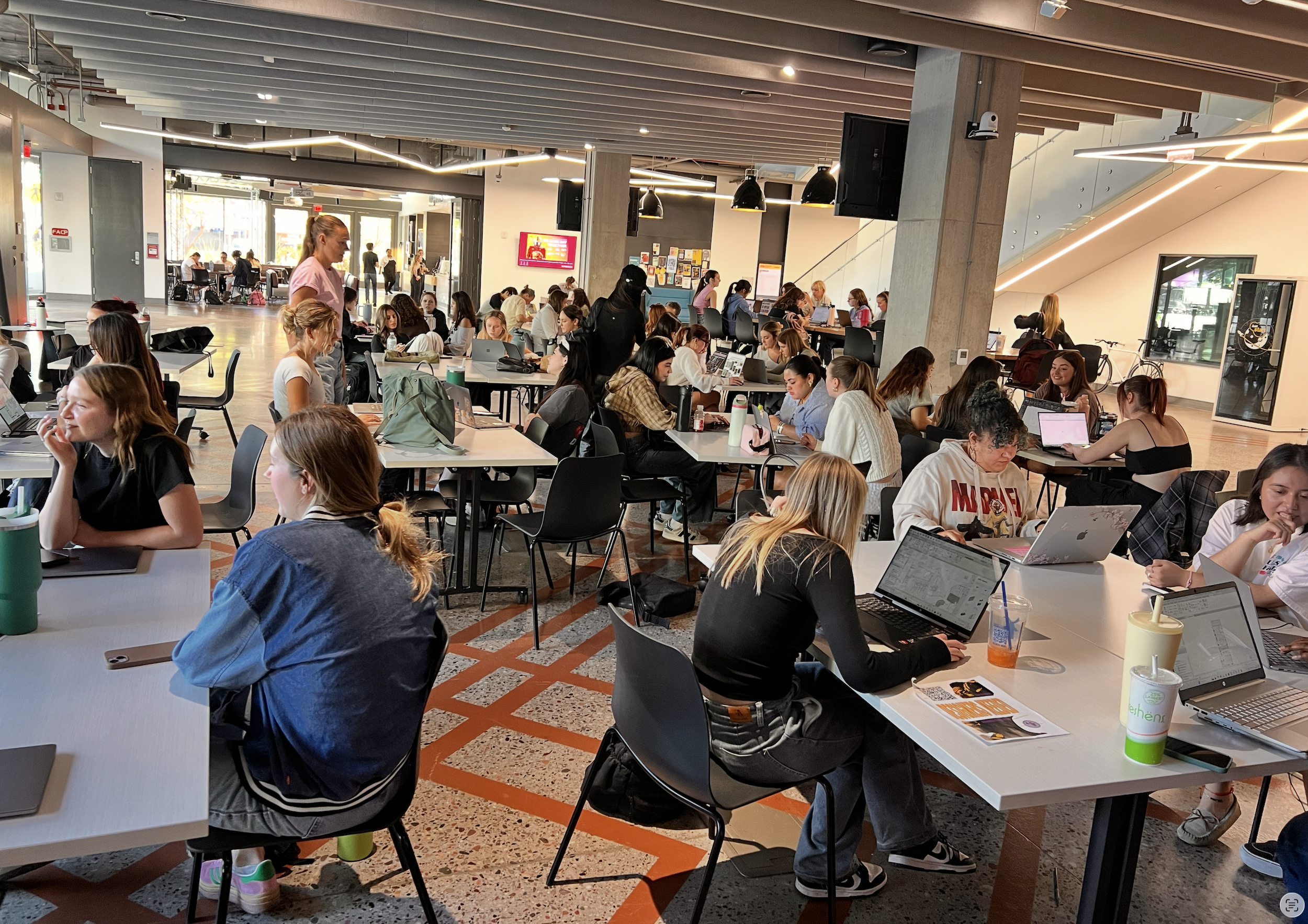 A busy, modern coworking space filled with people working on laptops at white tables, some talking on the phone, with large windows, a staircase, and decorative lighting fixtures.