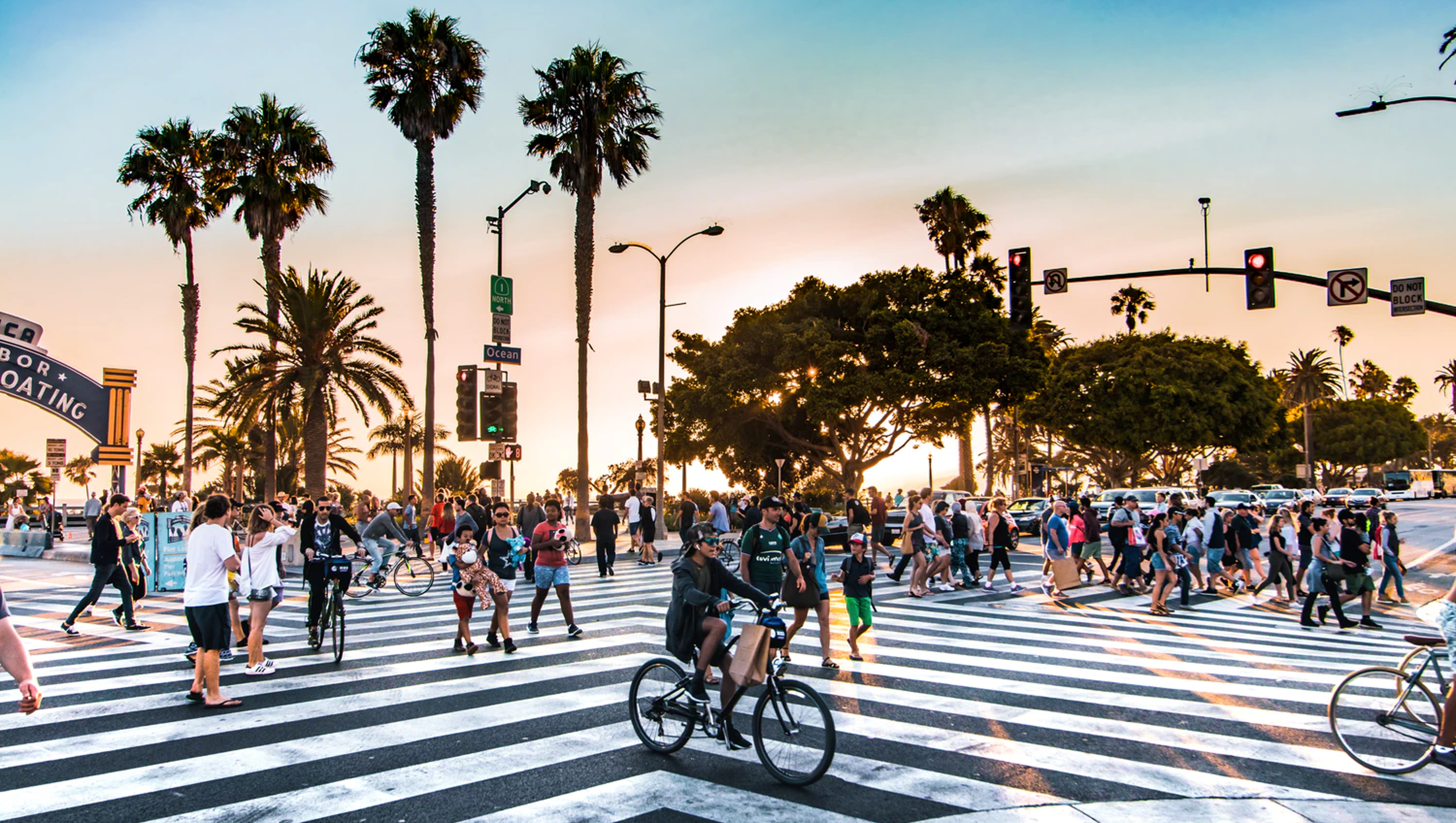 Crowd of people crossing a busy street at sunset, with palm trees and traffic lights in the background, in a lively urban area.