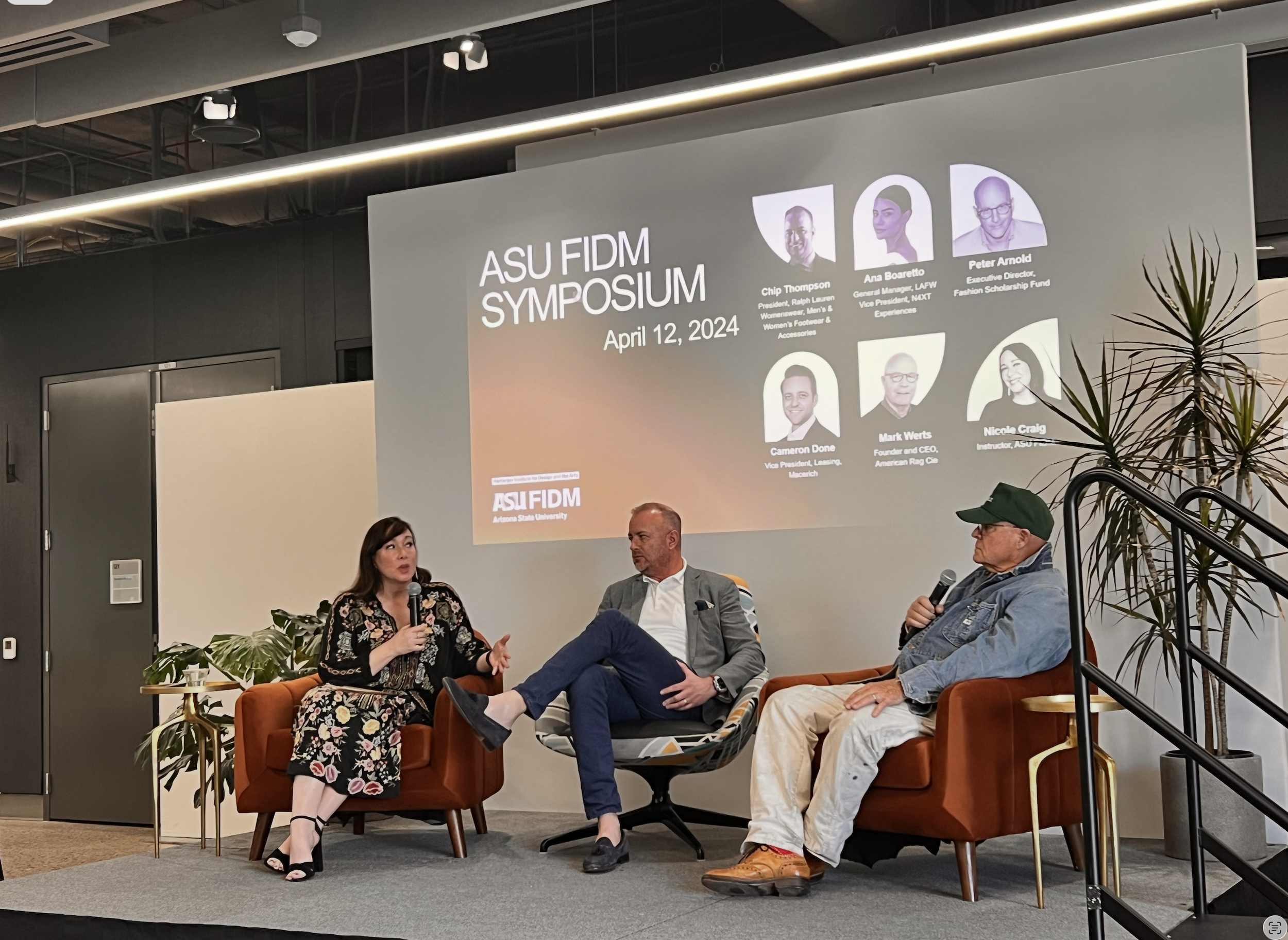 Panel discussion at the ASU FIDM Symposium on April 12, 2024, with four speakers seated on a stage, a large screen displaying event details, and a modern conference room setting.