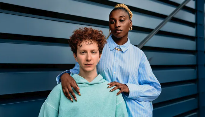 Two women standing outdoors against a modern building wall, one with curly red hair wearing a mint green hoodie, the other with dark hair in a bun wearing a light blue striped shirt.