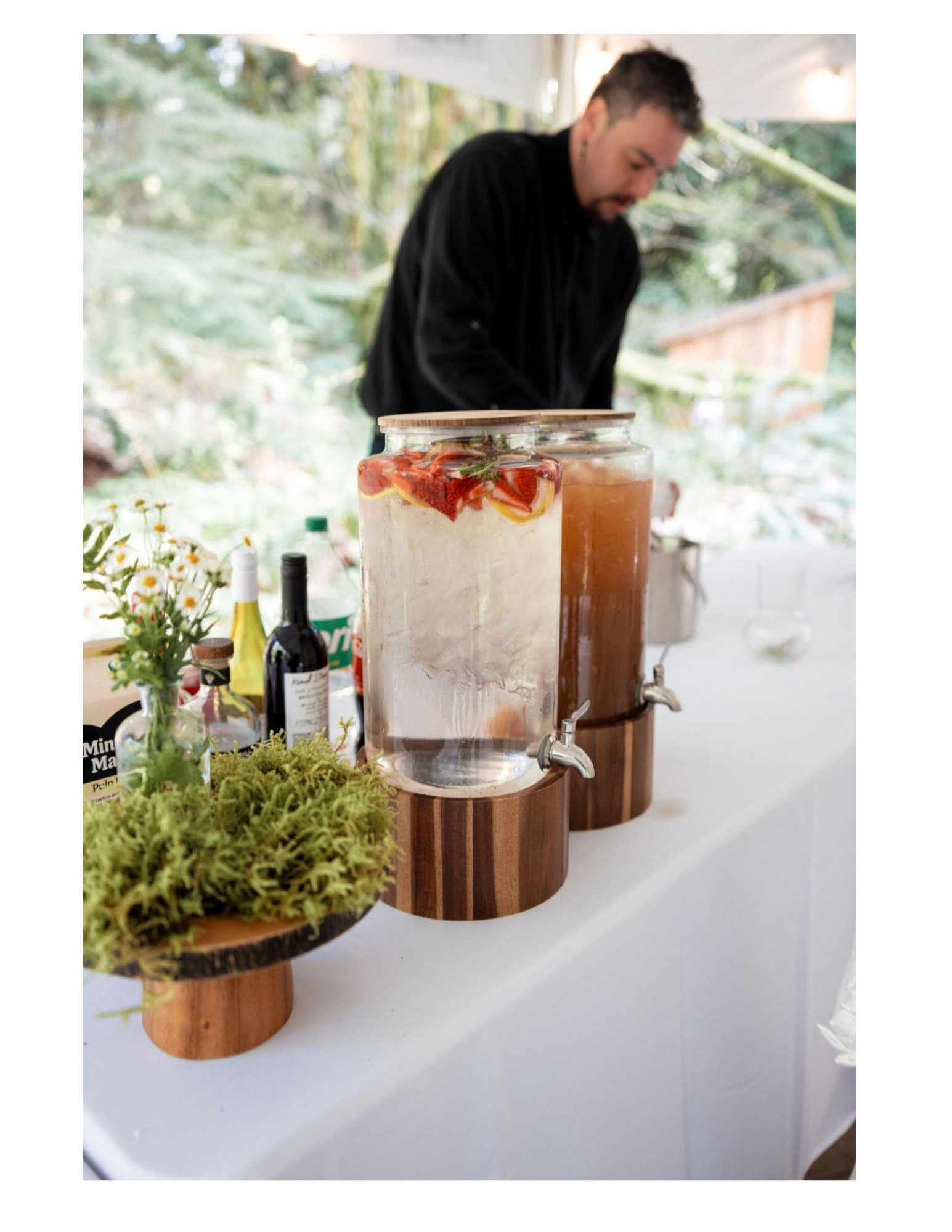A man in black shirt standing behind a table with large glass beverage dispensers filled with strawberry and lemon infused water and iced tea, decorated with flowers and greenery, outdoors with trees in the background.