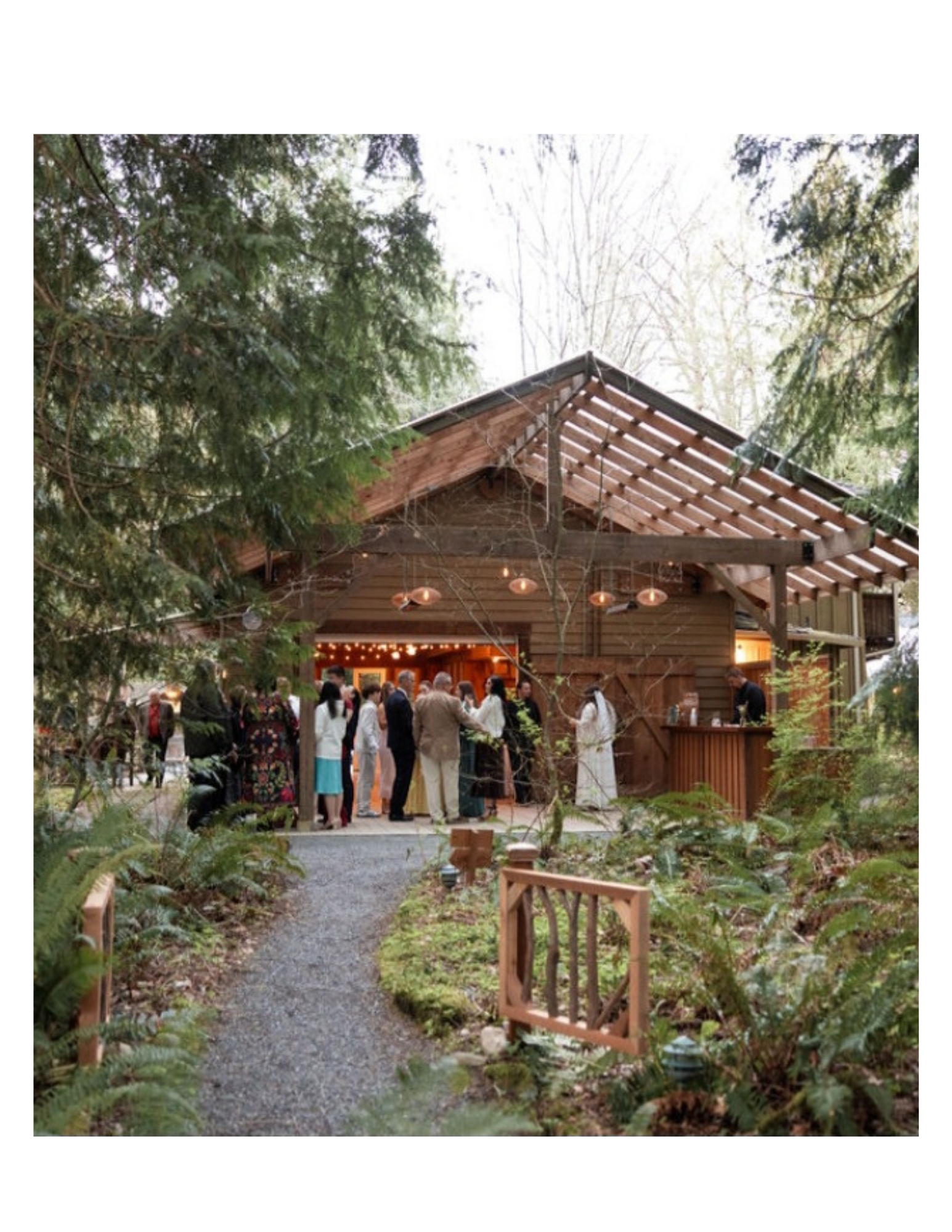 People gathered at an outdoor event in a wooded area with a wooden barn-like structure, string lights, and lush green trees.