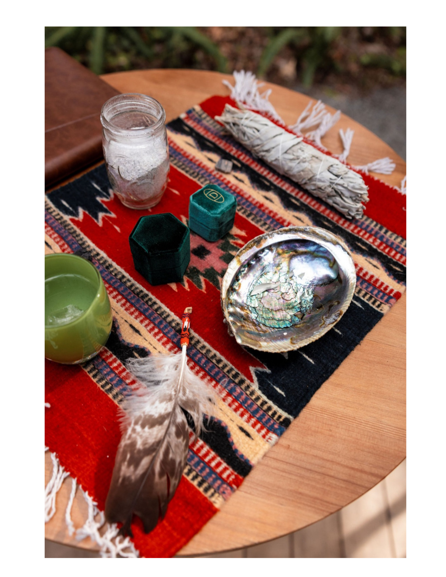A table set with a piece of red and black patterned textile, a green candle in a glass holder, a pair of green velvet jewelry boxes, a mussel shell filled with pearls, and a feather quill pen. In the background, there is a clear glass jar filled with white powder and an item wrapped in white string.