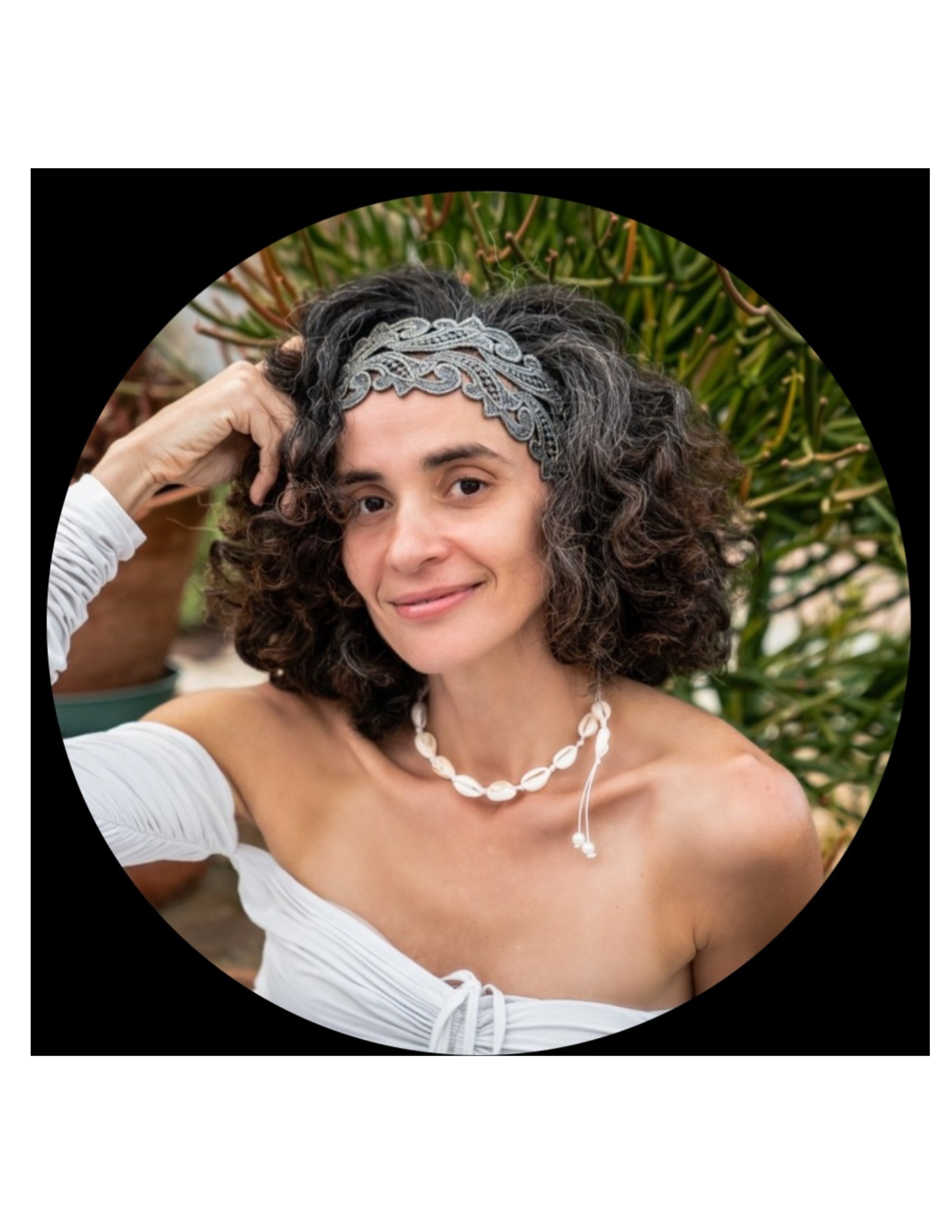 A woman with curly hair and a decorative headband, wearing a white off-shoulder top and a shell necklace, smiling outdoors with green foliage in the background.