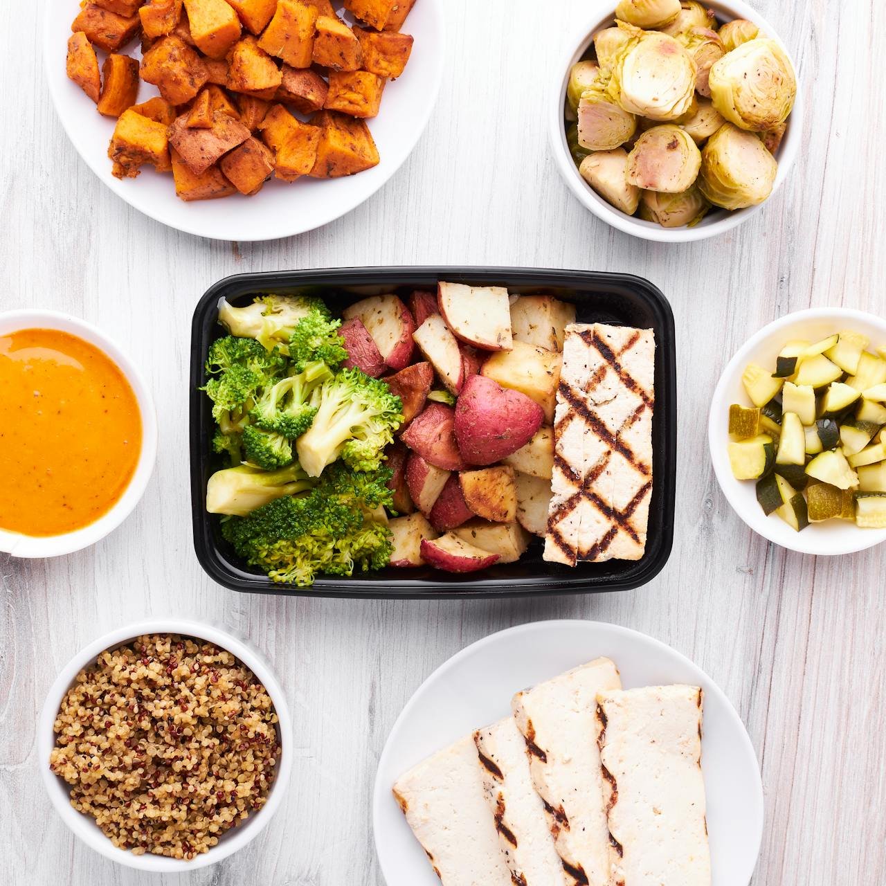 An assortment of cooked vegetables, grains, and grilled meats arranged on white and black dishes, including roasted sweet potatoes, grilled halloumi cheese, broccoli, sweet potatoes, and other vegetables, with side sauces and dips.