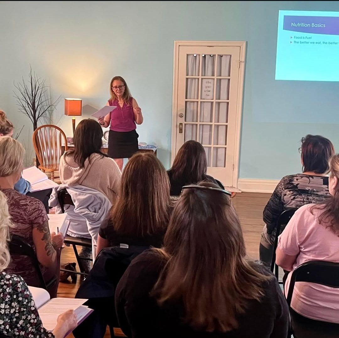 A woman giving a presentation to a group of women seated in a room with light blue walls. The presentation slide on nutrition basics is projected on the wall.