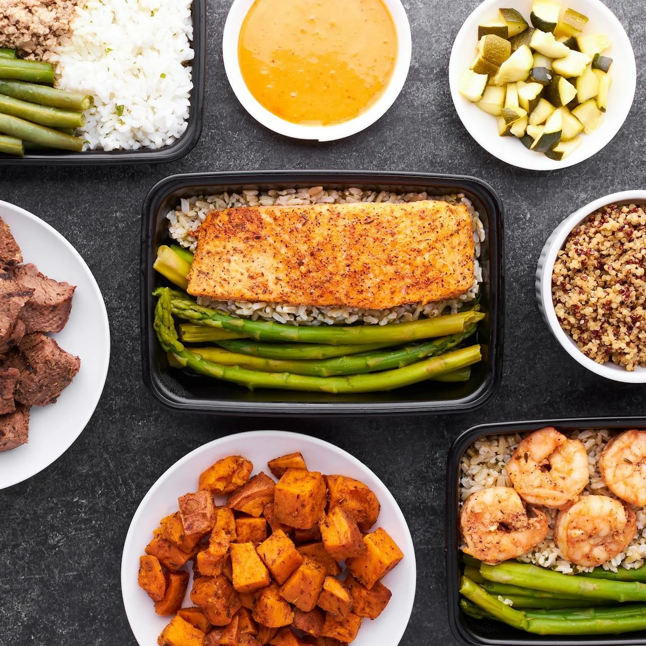 A variety of prepared dishes including salmon with asparagus, cooked shrimp with rice, roasted sweet potatoes, chopped pickles, fresh green beans and rice, and a bowl of quinoa, all arranged on a dark countertop.
