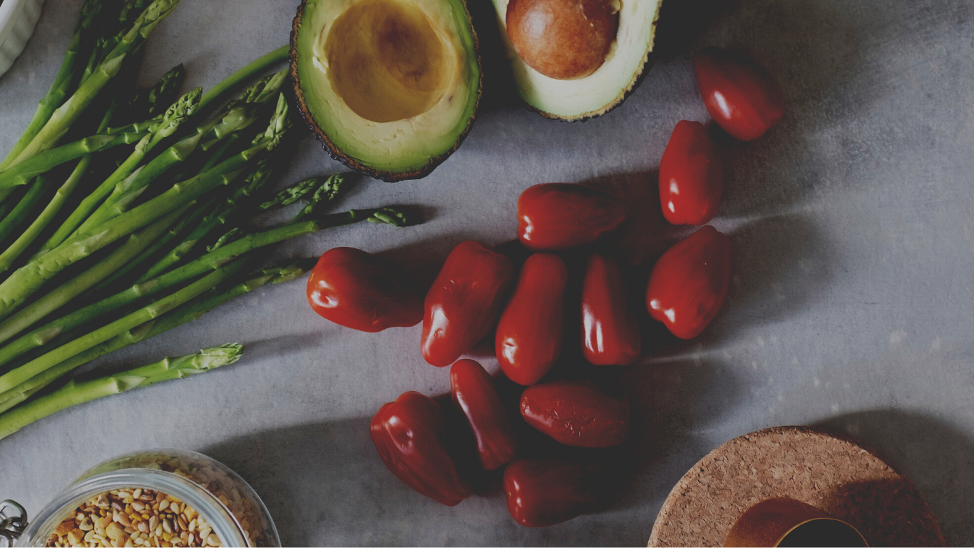 Fresh vegetables including a bunch of green asparagus, halved avocados, grape tomatoes, and a jar of chopped nuts on a gray surface.