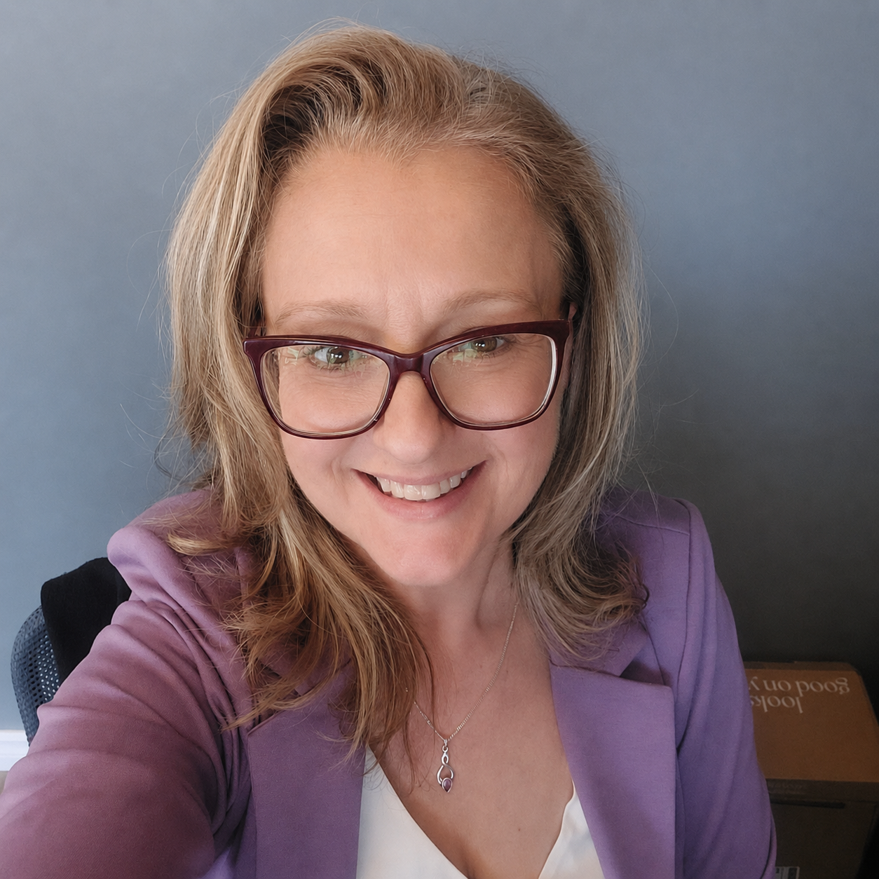 A woman with shoulder-length blonde hair and glasses smiling at the camera, wearing a purple blazer and a white top, with a book partially visible in the background.