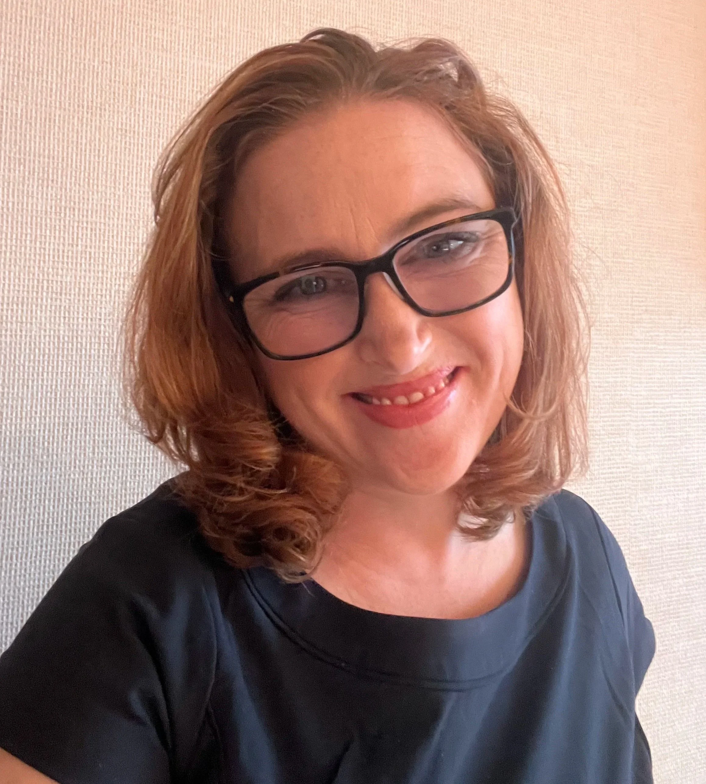 Close-up of a woman with light brown, curly hair and black glasses, smiling, wearing a black top, with a textured beige wall in the background.