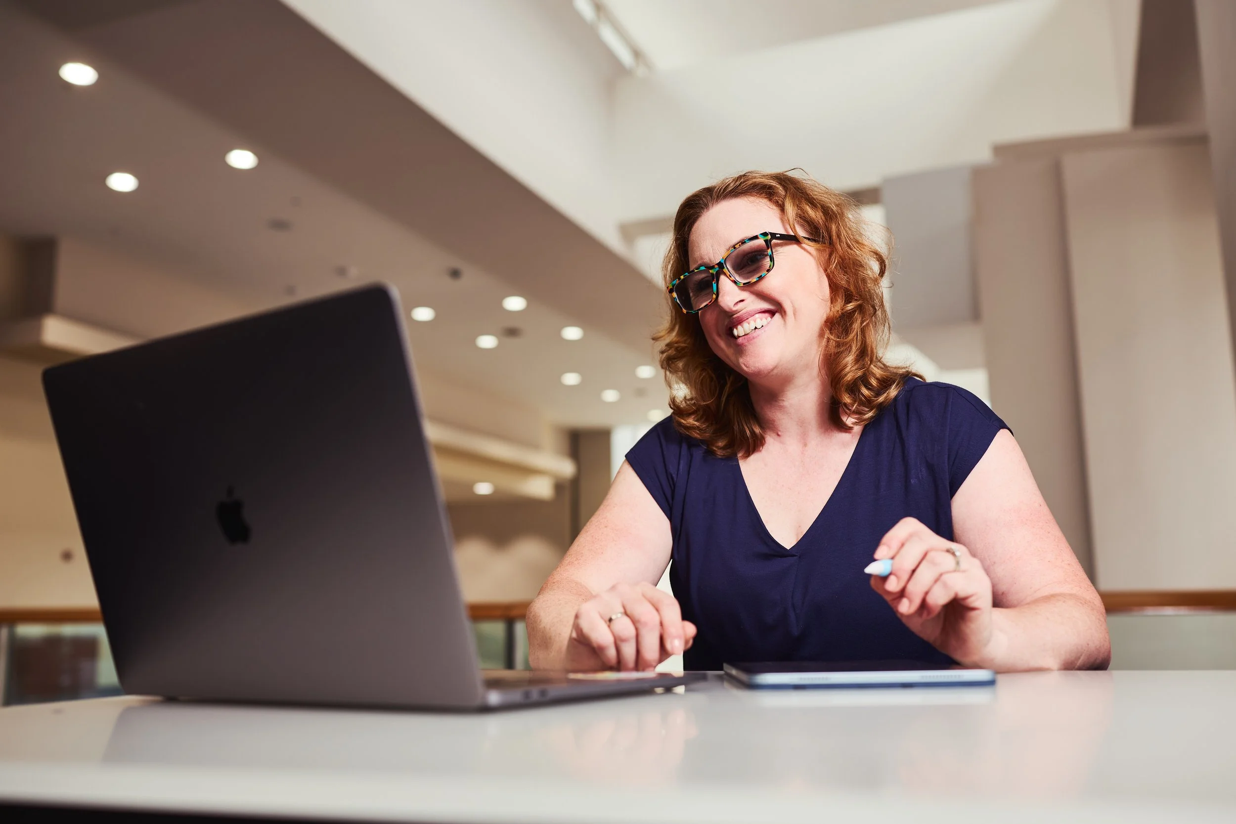 Dr. Melanie Barham is smiling and looking at her laptop while holding a pen, seated at a table in a well-lit indoor setting.