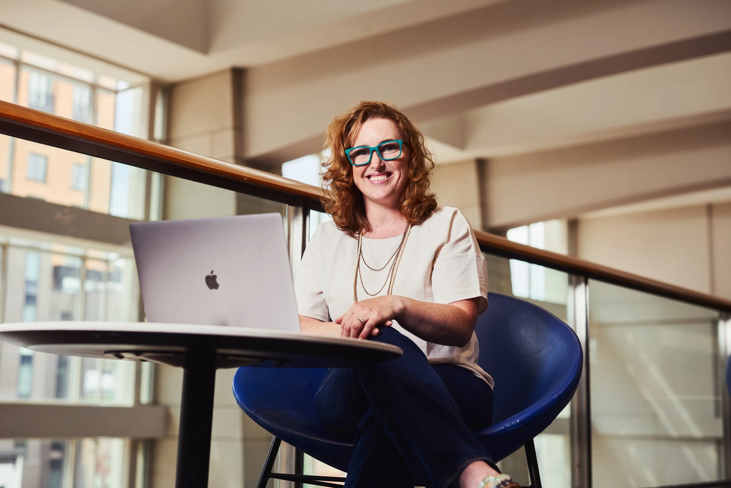 Dr. Melanie Barham is seated at a table with a silver Apple MacBook laptop in a modern building with large windows.