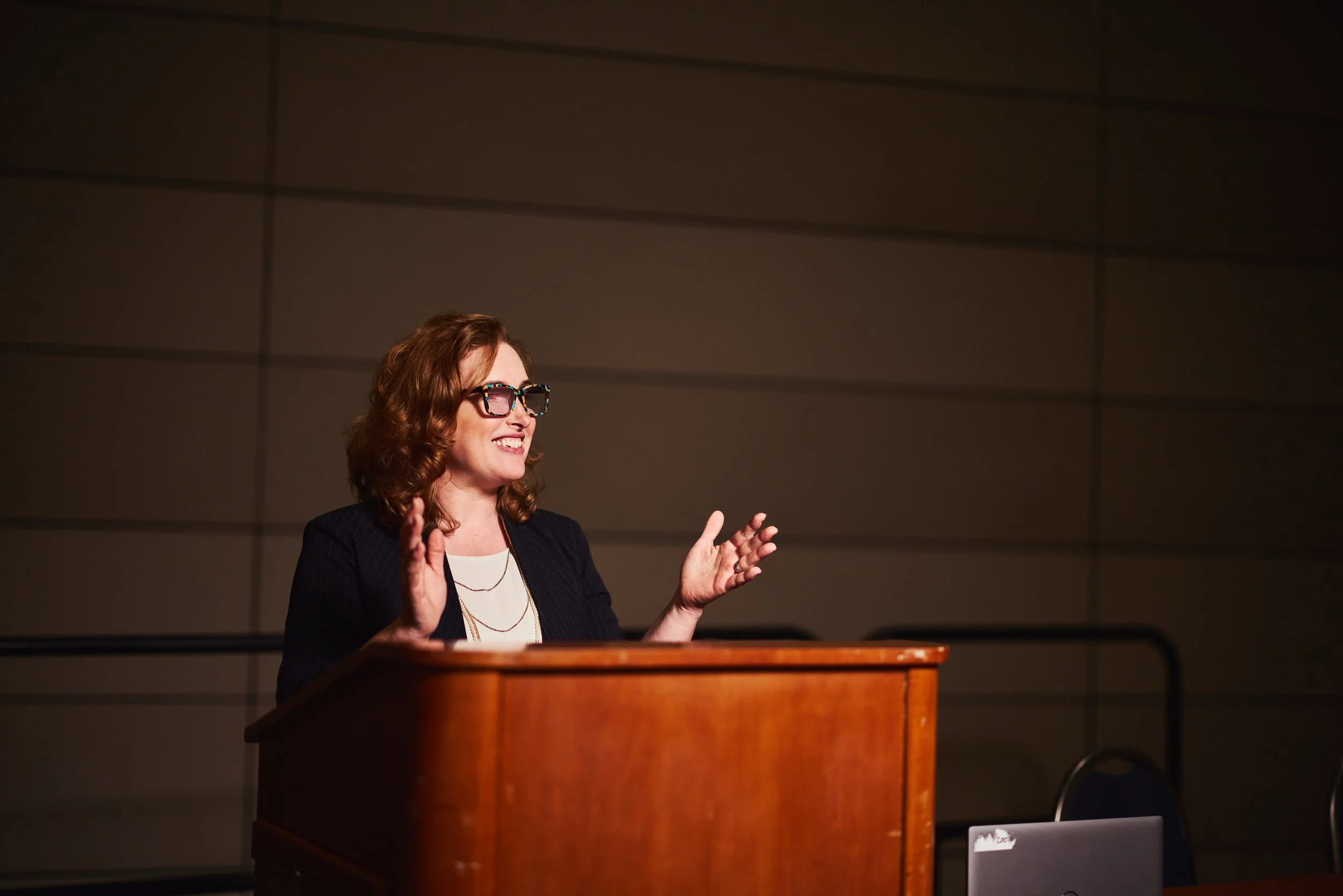 Dr. Melanie Barham is speaking at a podium, gesturing with her hands in front of a plain wall.