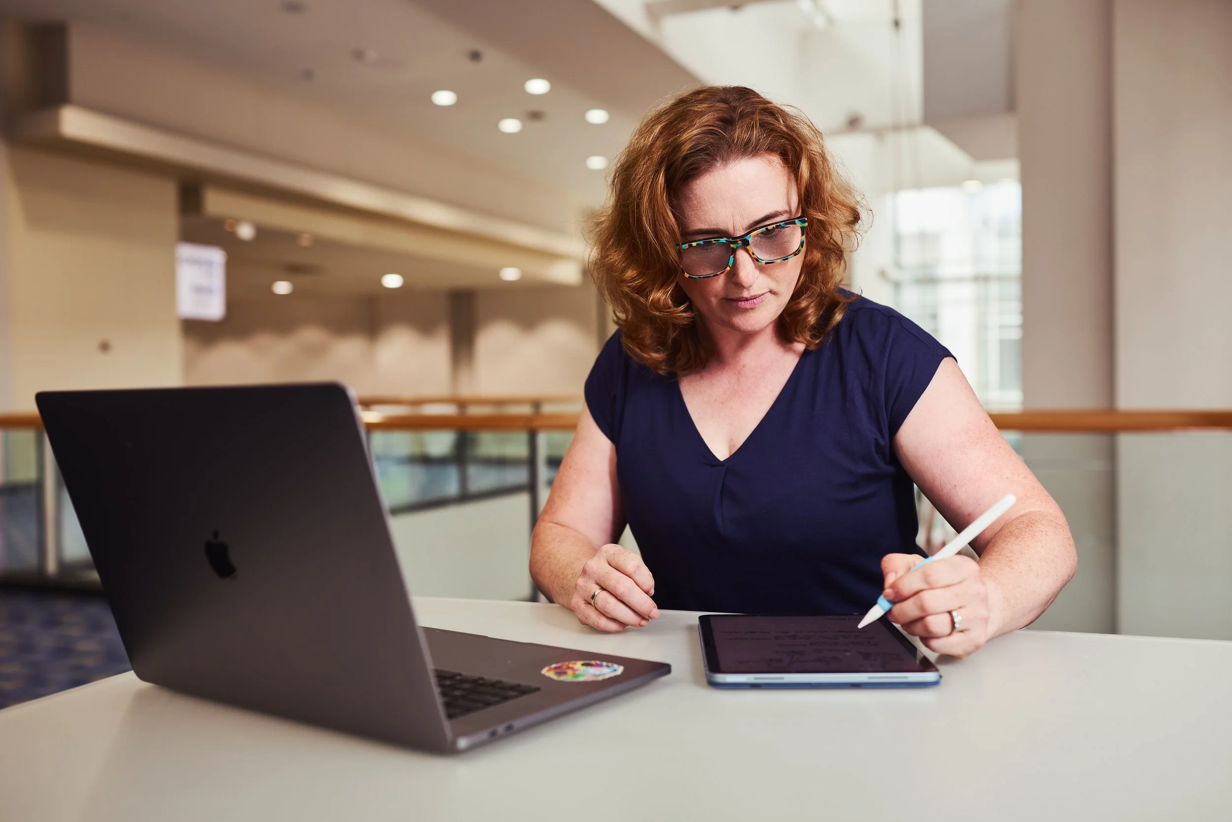 Dr. Melanie Barham is sitting at a table, working with a stylus on a digital tablet, with a laptop open in front of her in a modern, well-lit space.