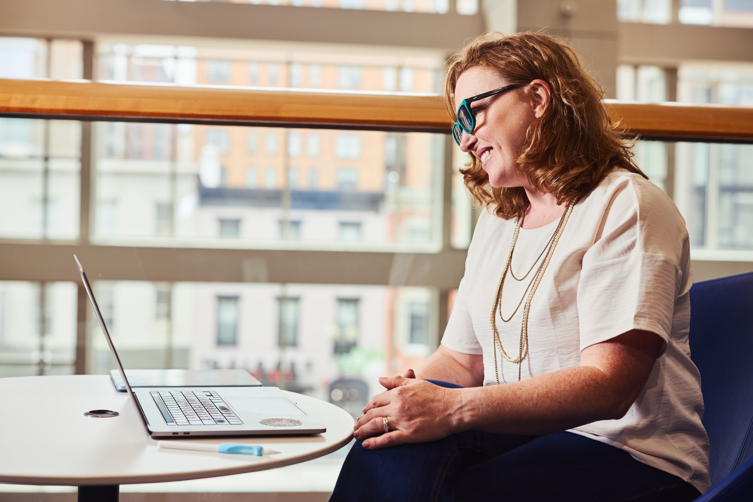 Dr. Melanie Barham is sitting at a table with a laptop in front of her, smiling while working indoors with large windows in the background.