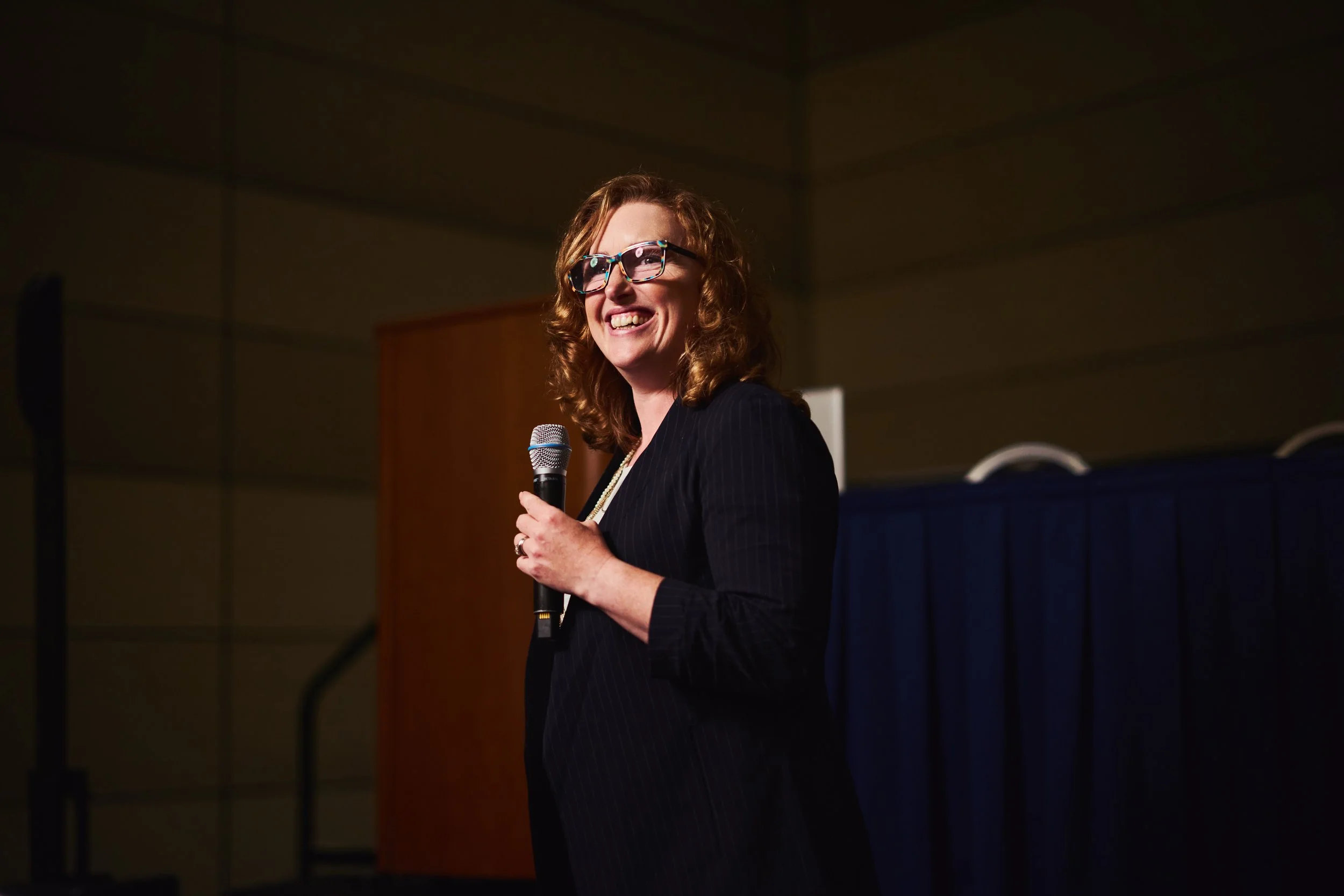Woman with curly hair and glasses smiling while holding a microphone in a room with wood-paneled walls.