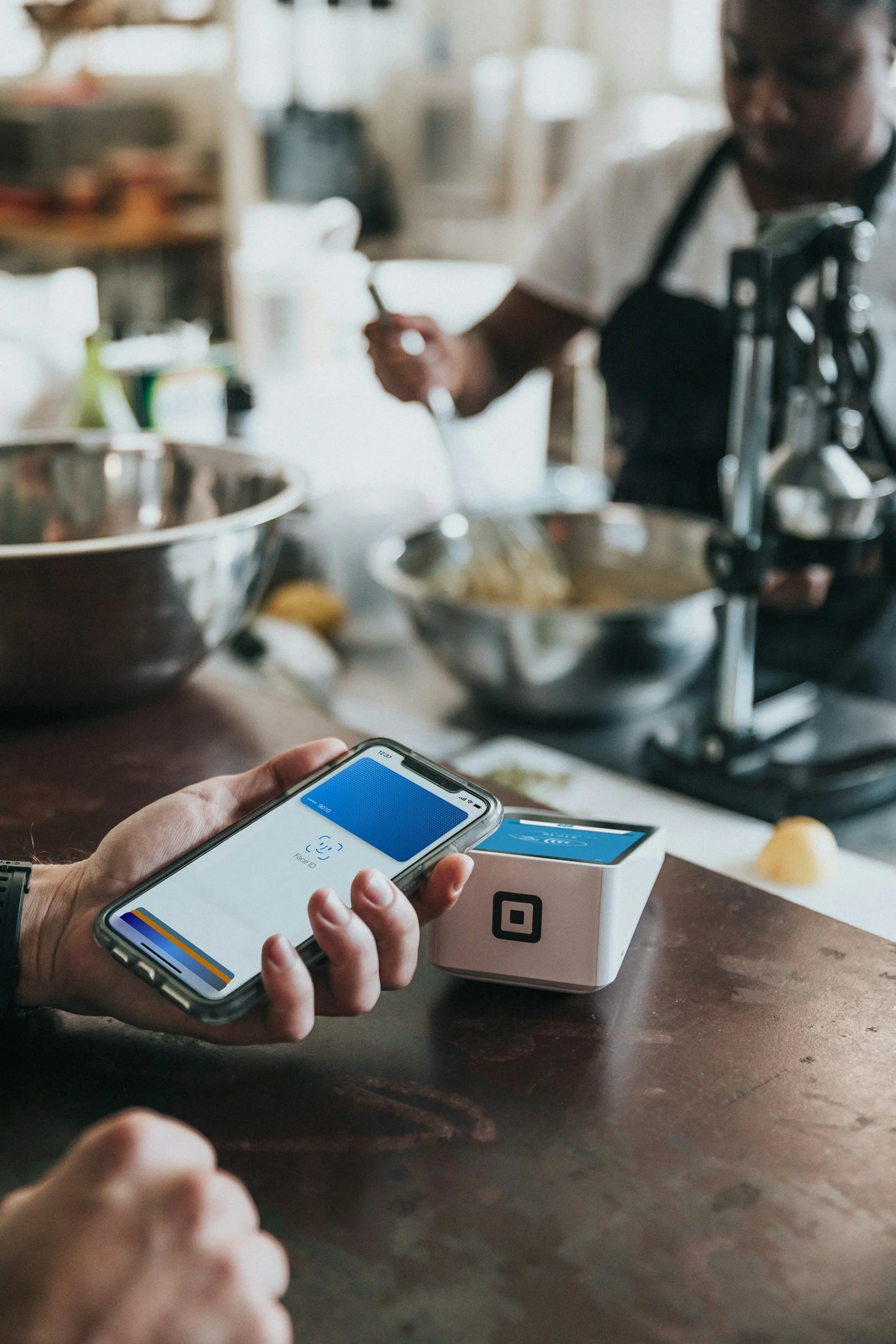 Person using a smartphone with facial recognition app in a kitchen, with a payment terminal on the counter.