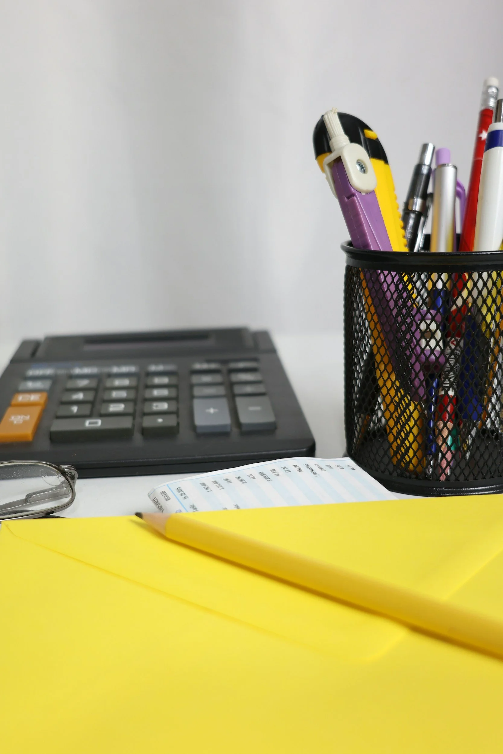 Office desk with a black container holding pens, markers, and a tape dispenser, a calculator, glasses, a yellow notepad, and a paper with printed text on a white surface.