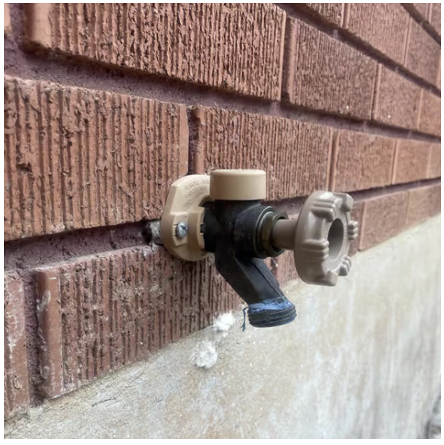 A garden hose bibb or outdoor water faucet attached to a brick wall, with a black hose and a beige valve handle.