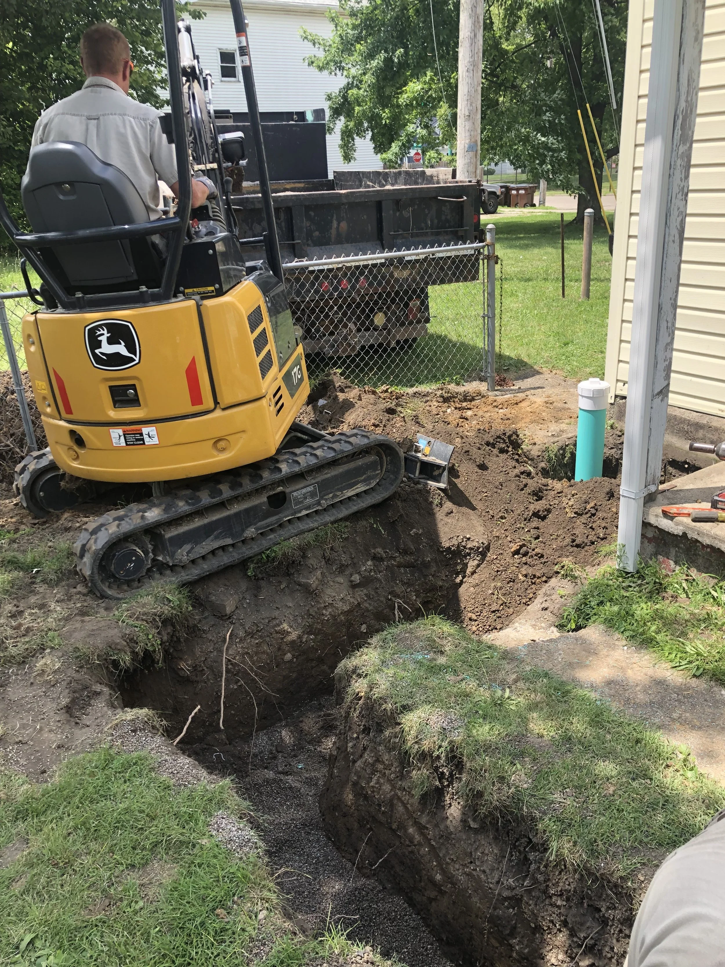 A construction worker operating a small excavator digging a trench next to a house. The trench is filled with black gravel or soil, and a pipe is visible on the right side of the image. There is a chain-link fence and a green yard with trees and neighboring houses in the background.