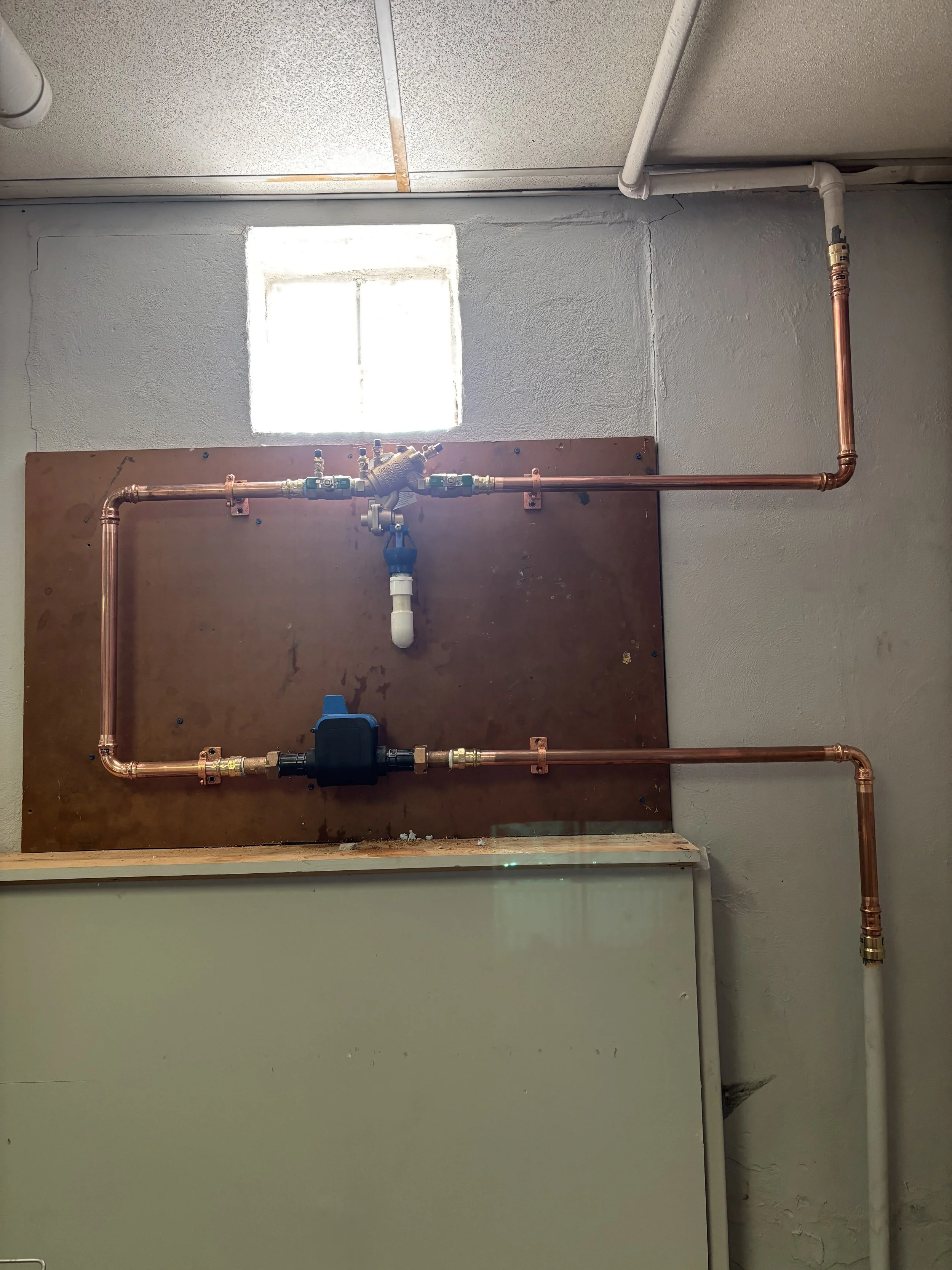 Interior view of a wall with copper pipes, valve, and piping connections, with a small window near the ceiling illuminating the space.