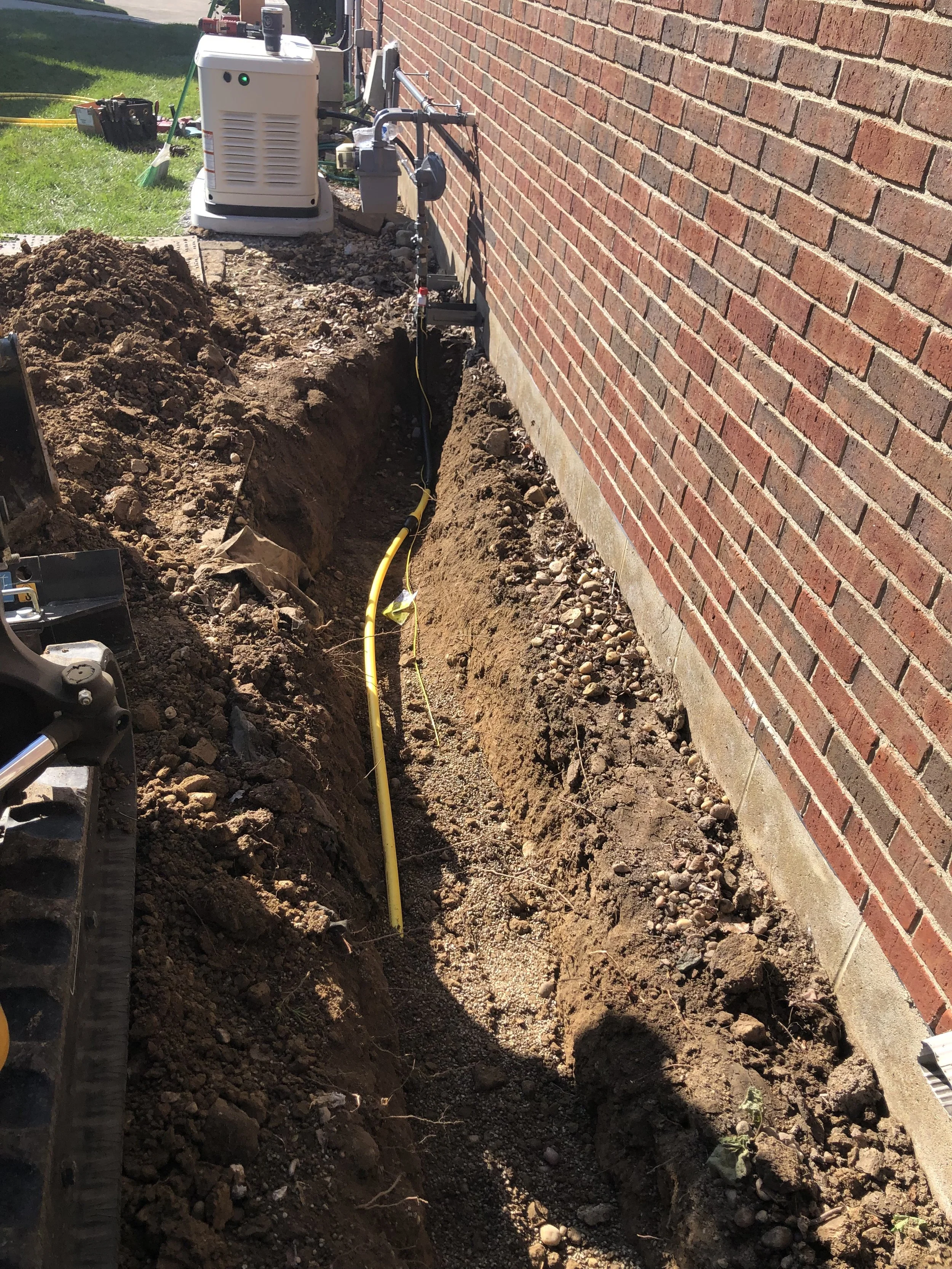 A trench dug alongside a red brick wall, with yellow and black tubing or piping running through it. Construction equipment and tools are visible in the background on a grassy area.