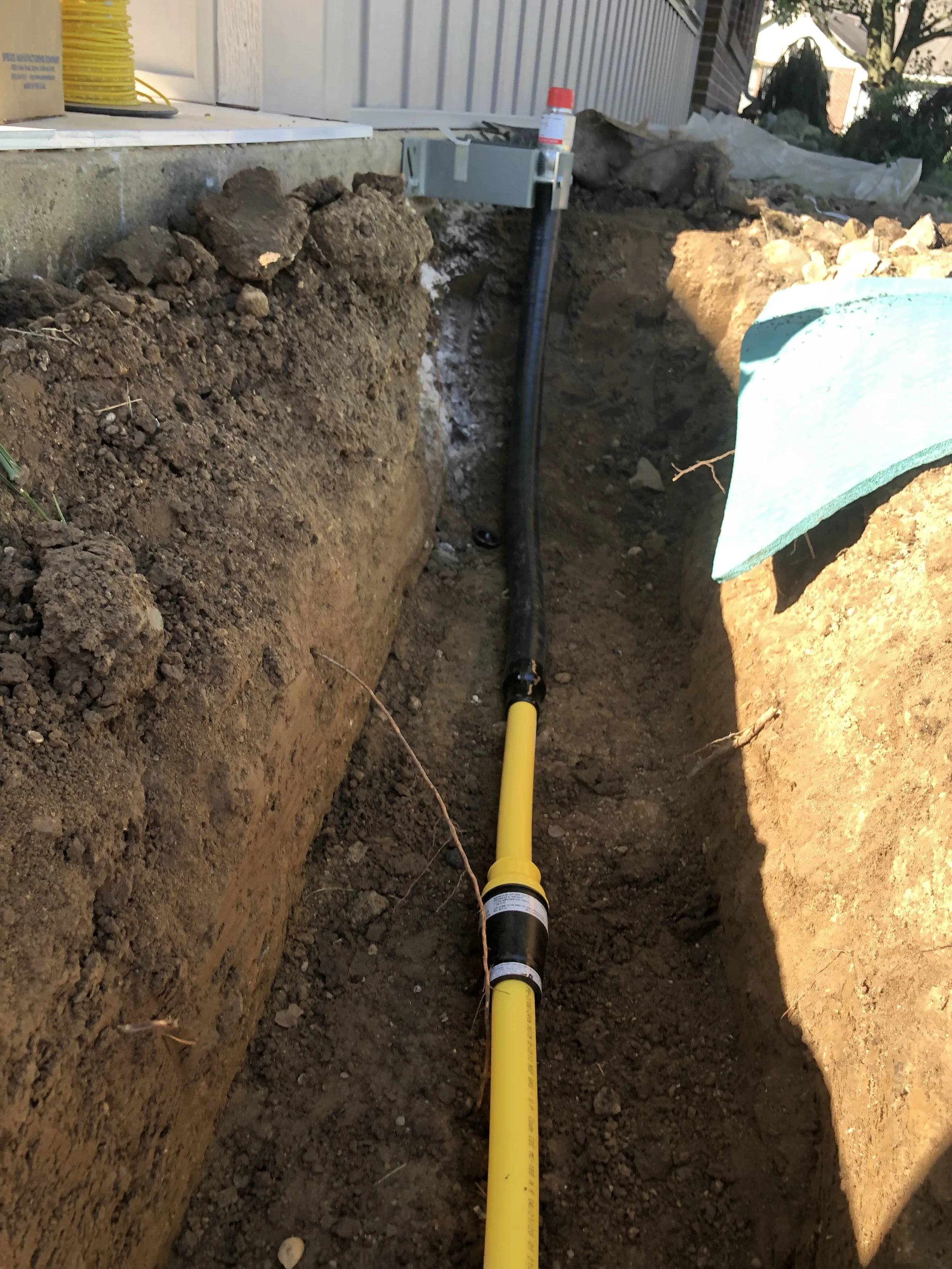 Yellow underground pipeline in a construction trench, with a black and yellow valve attached to it, near a house foundation.