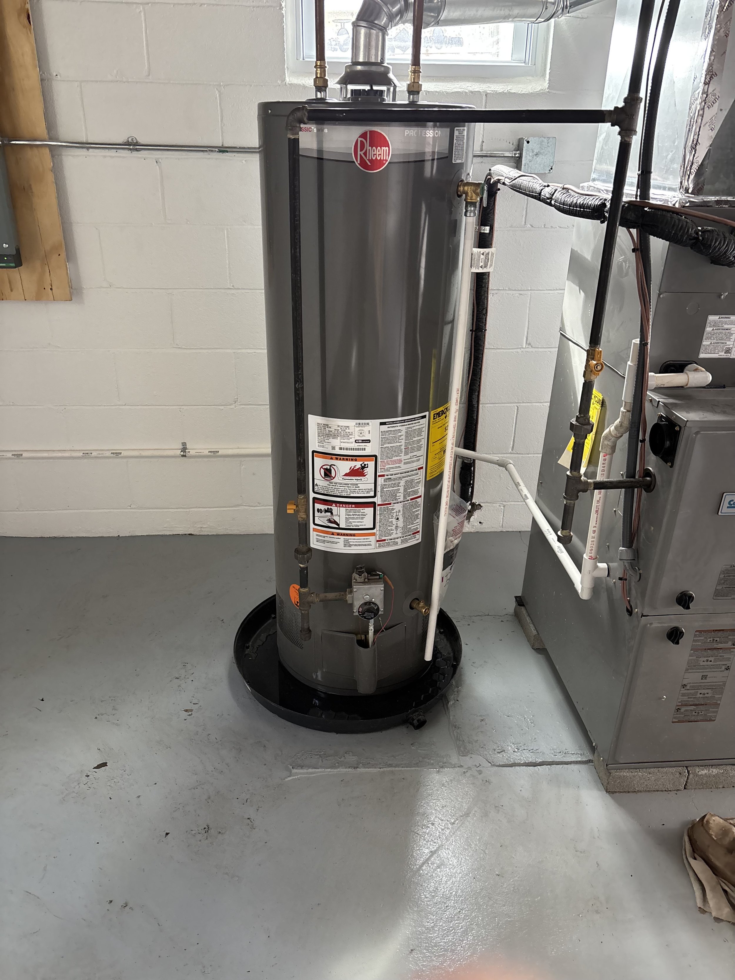 A water heater tank in a utility room with pipes and a window in the background. The tank has a Rheem logo and safety labels on it.