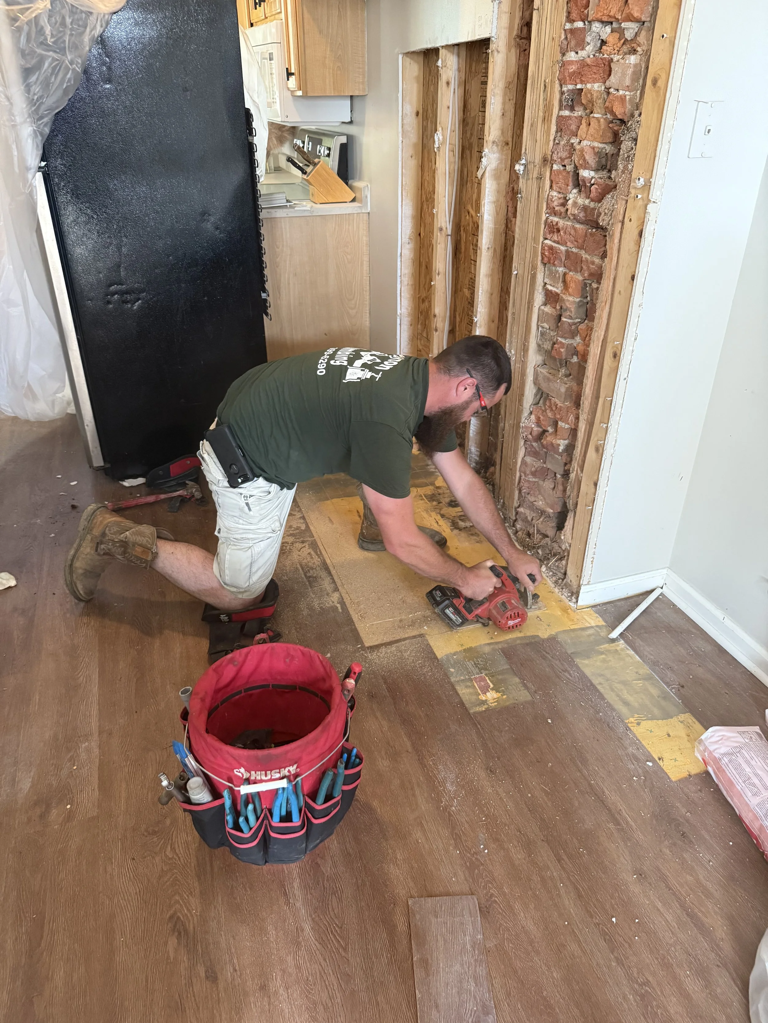 A man with a beard and glasses kneeling on the floor, working on a wall with exposed brick and wood framing. He is using a power tool and is surrounded by tools in a red Husky tool bag. The room appears to be under renovation with some flooring removed.