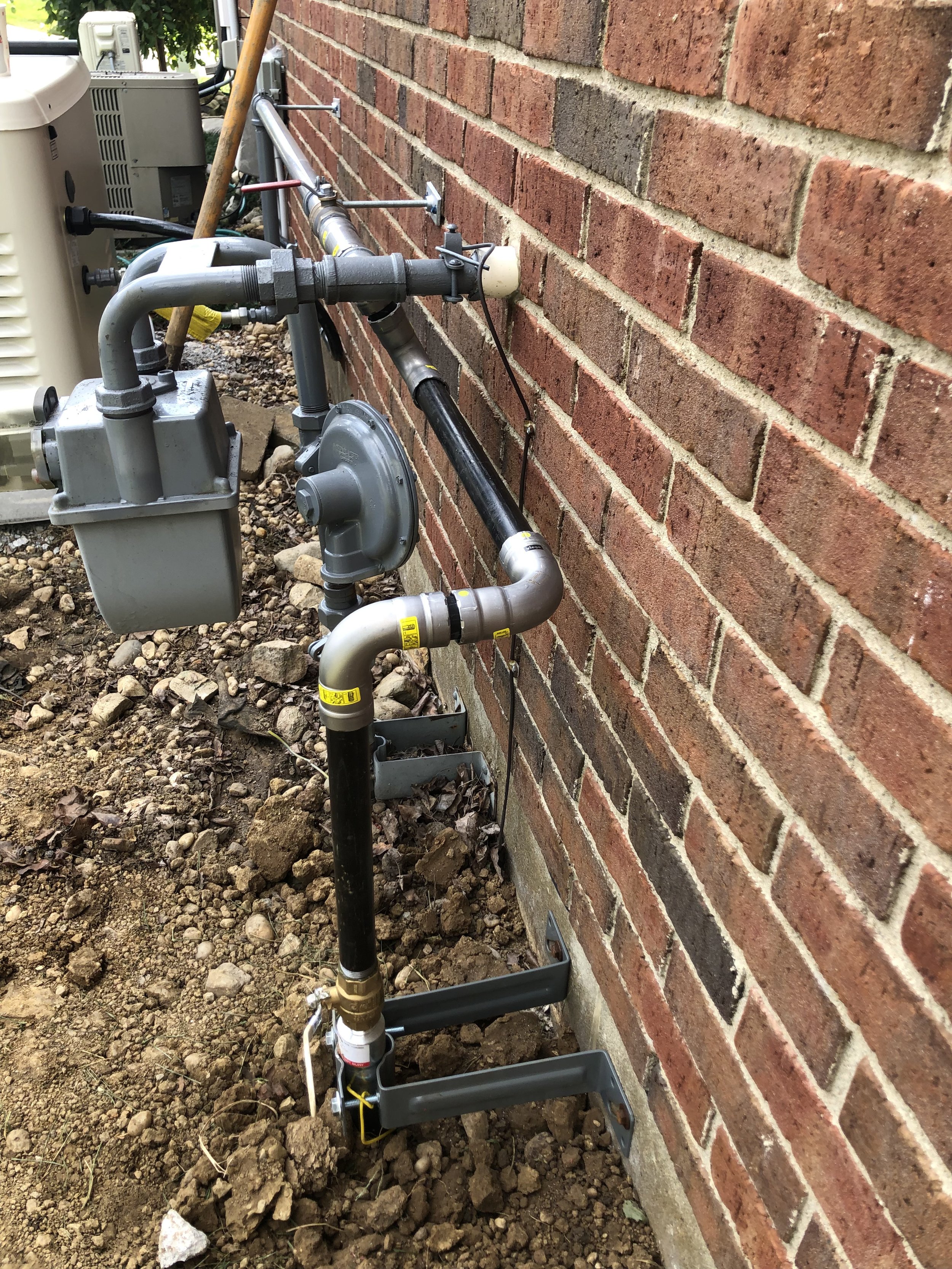 Outdoor view of plumbing and gas line setup against a red brick wall, with pipes, valves, and electrical components, surrounded by dirt and rocks.