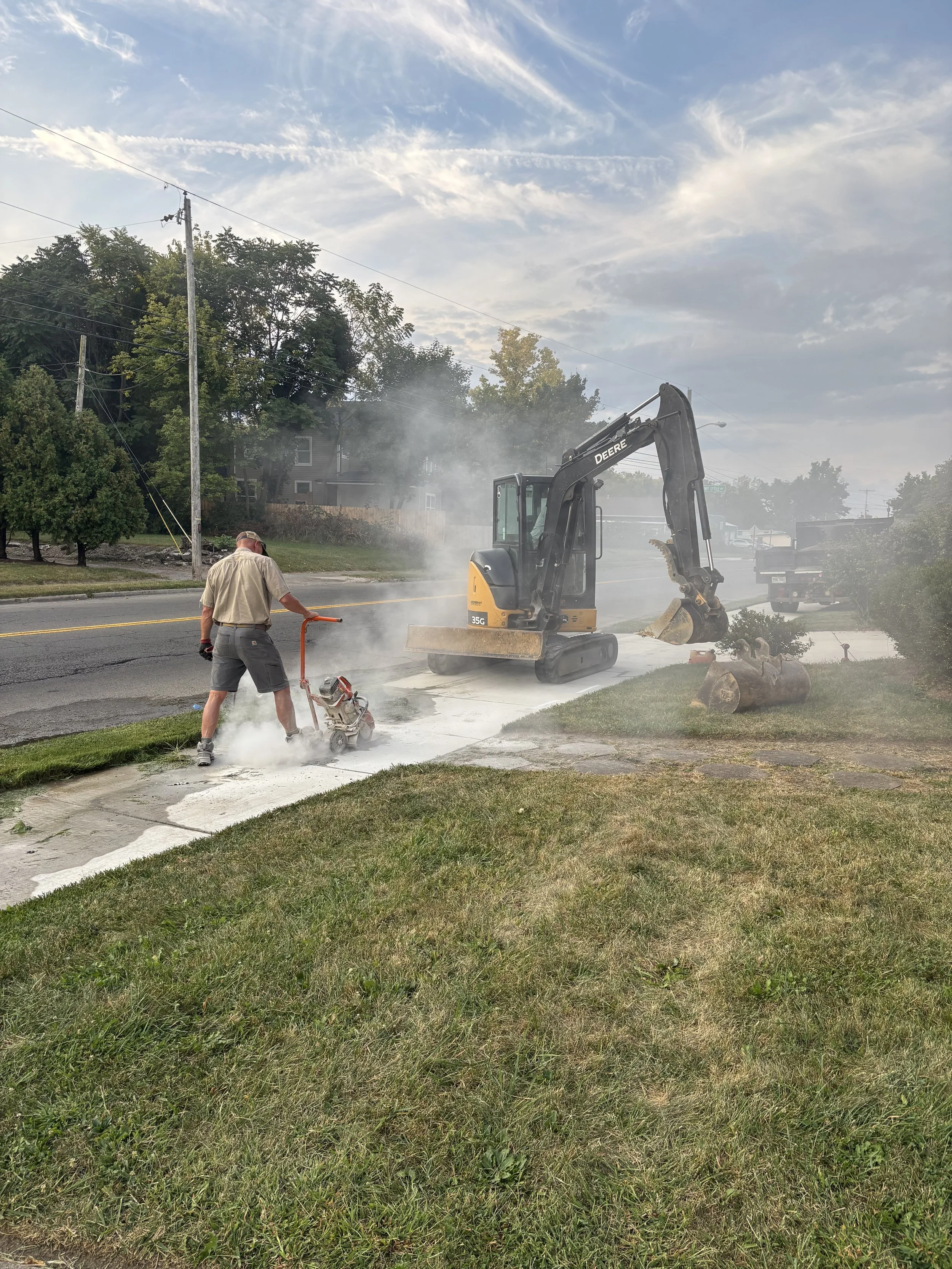 A man operating a small excavator parked next to a street while another man uses a concrete saw on the sidewalk during daylight hours, with trees, houses, and clouds in the background.