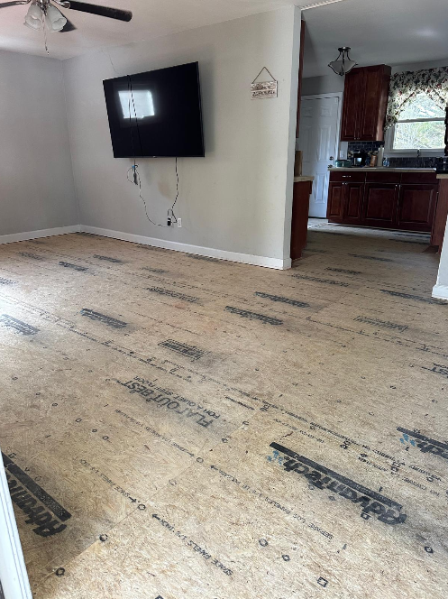 Sala vacía con piso en proceso de instalación, pantalla plana montada en la pared, y cocina visible en el fondo.