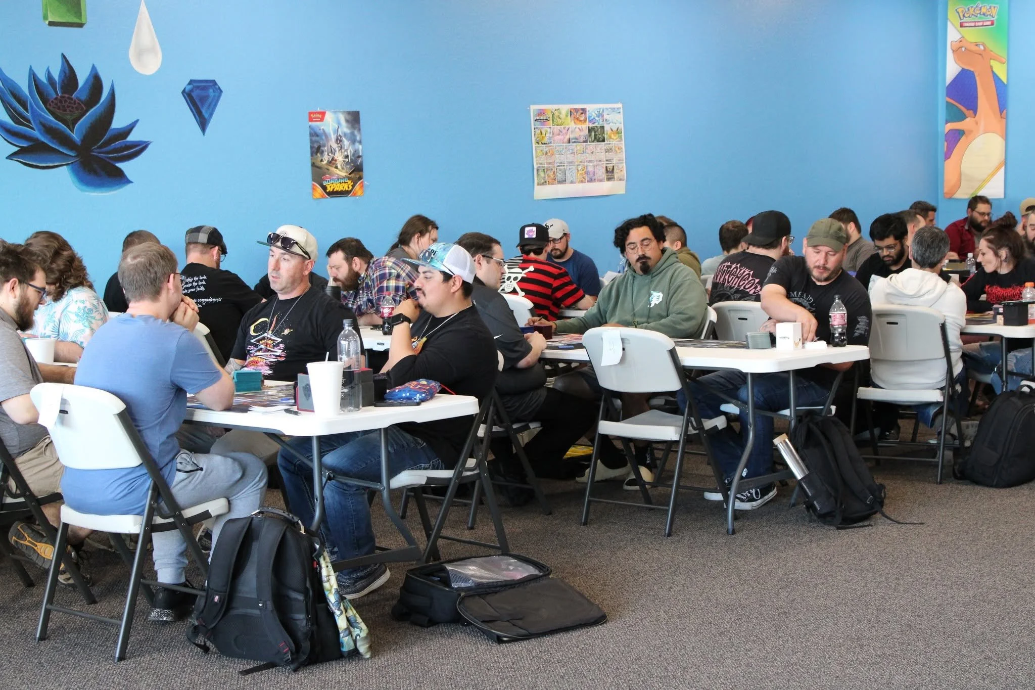 A group of people seated at tables in a room decorated with Pokémon posters and wall art. They appear to be playing games or engaged in a group activity.