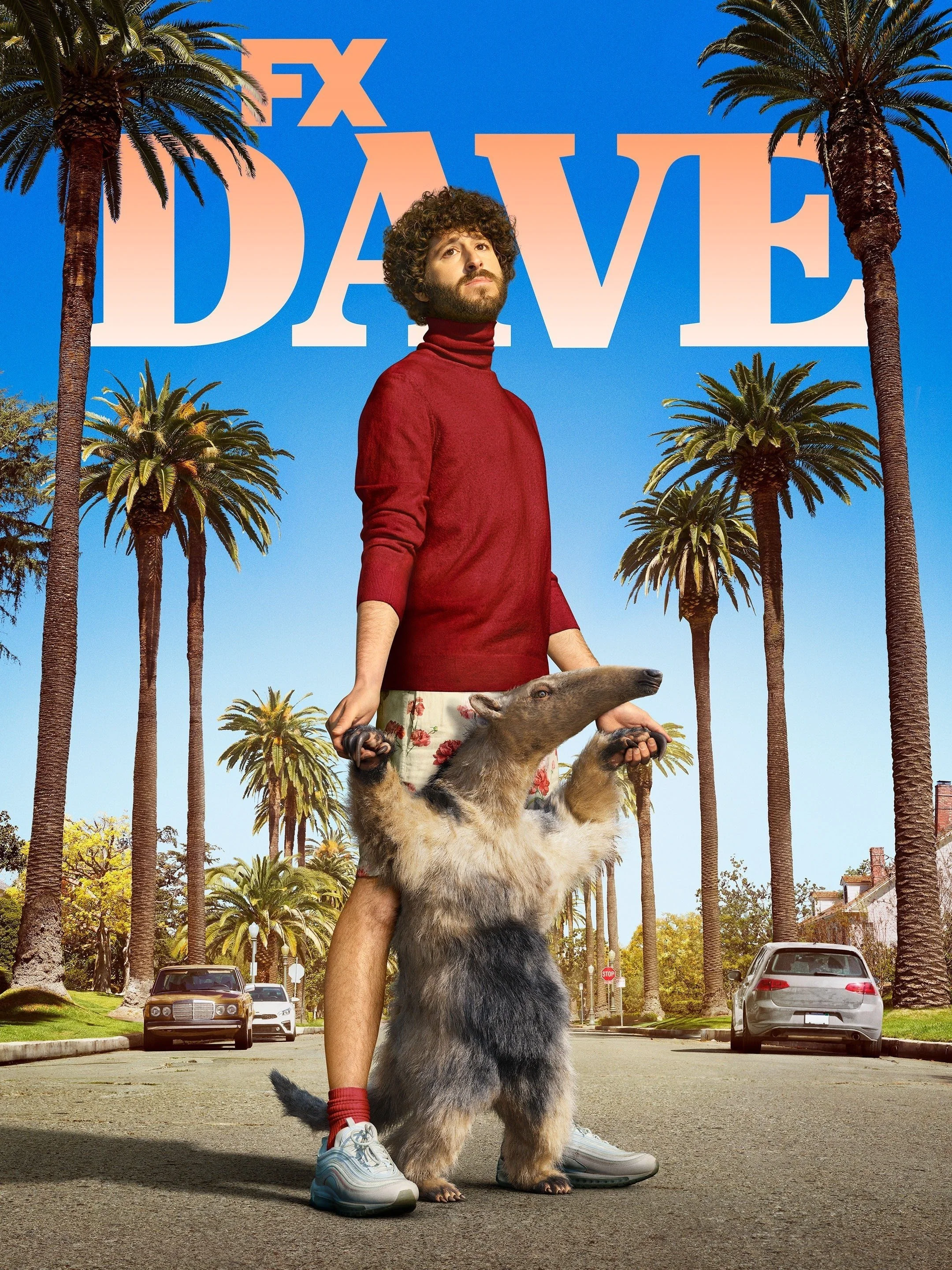 A man with curly hair and a beard stands on a street with palm trees, wearing a red turtleneck, floral shorts, and sneakers, holding a large German Shepherd puppy. Large 'LOVE' letters with the word 'DAVE' behind him are in the background.