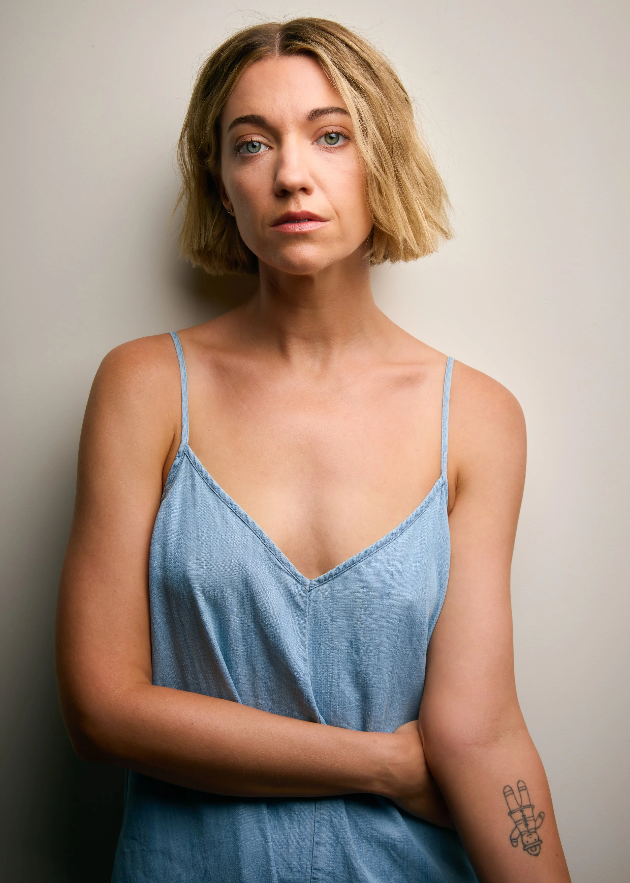 A woman with short blonde hair and blue eyes, wearing a light blue sleeveless top, standing against a plain off-white wall, with her arms crossed and a tattoo of a cartoonish hand giving a peace sign on her forearm.