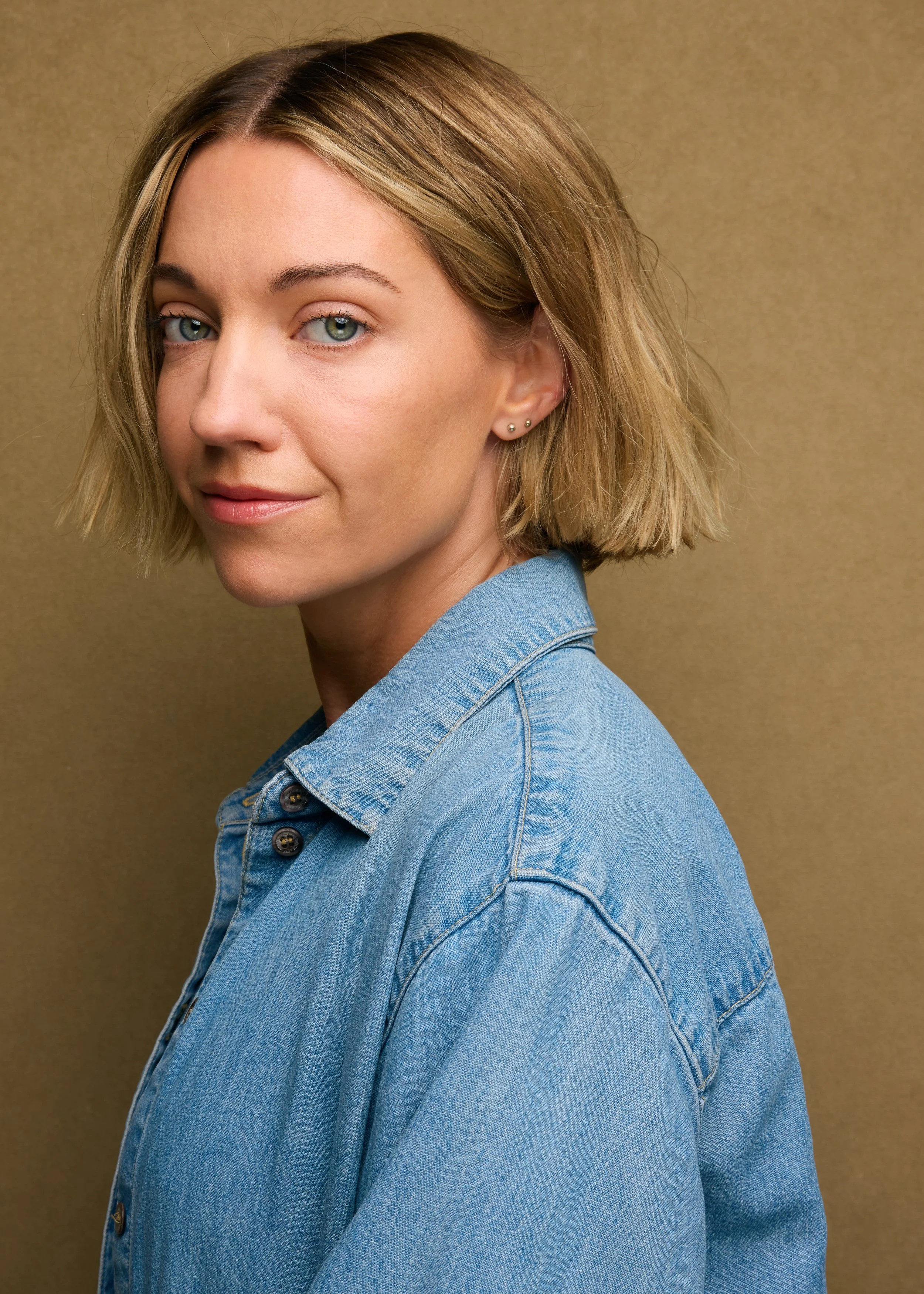 Close-up of a woman with short blonde hair, blue eyes, and fair skin, wearing a denim jacket, looking over her shoulder at the camera against a plain beige background.