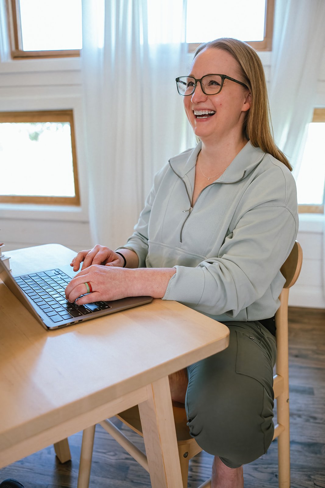 Founder of Root and Rise Consulting working on her laptop at a wooden table in a bright room with windows and curtains.