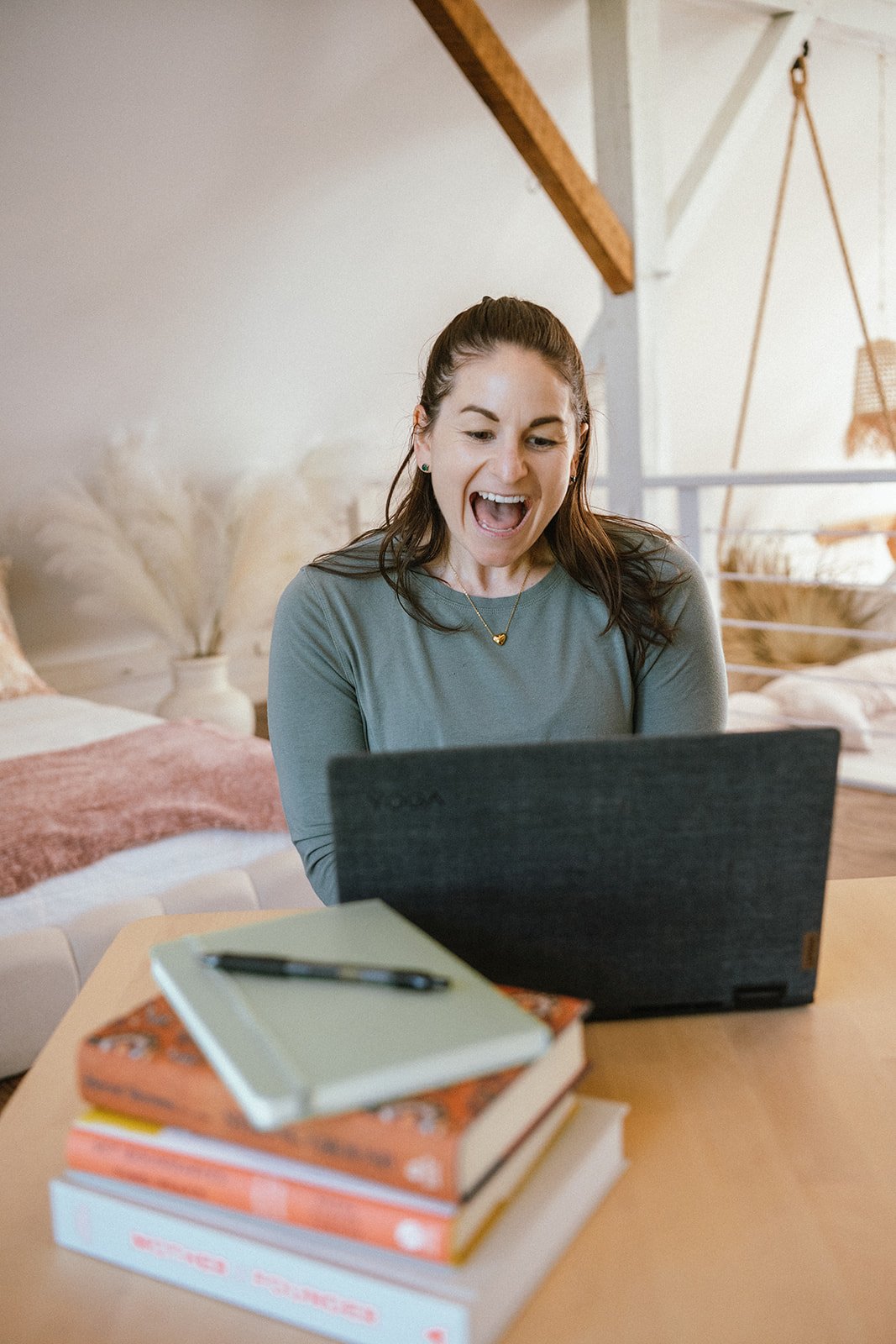 A woman with brown hair in a ponytail is excitedly looking at a laptop in front of her – Celebrating a win in her journey of work with Root & Rise Cosuling