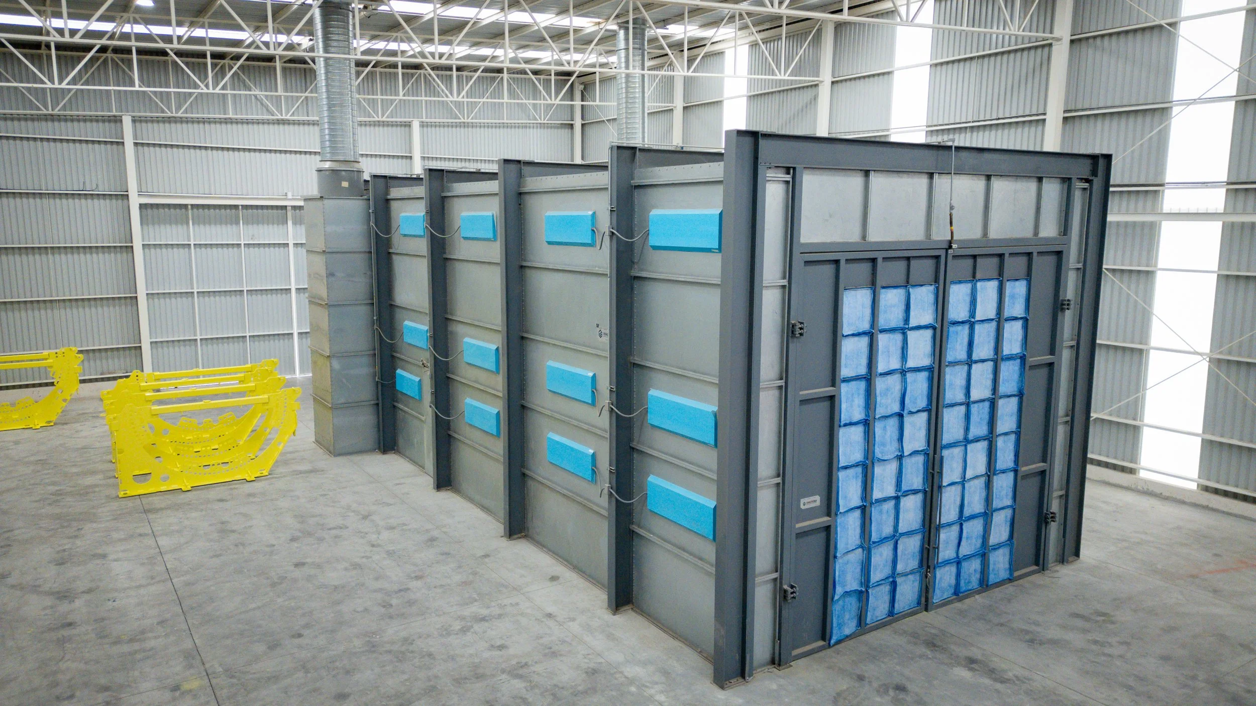 Infraestructura industrial con paredes metálicas y equipo de control o manipulación de líquidos, incluyendo una estructura grande con paneles azules y componentes metálicos, en un espacio amplio y techado.