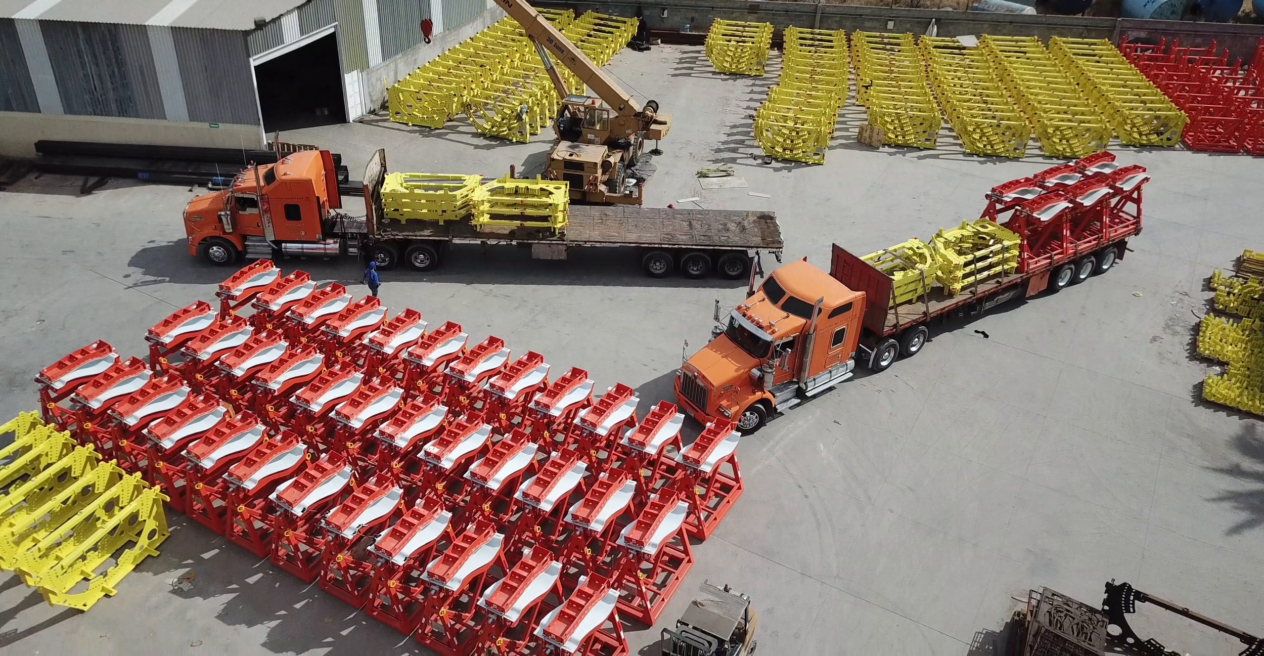 Imagen que muestra un almacén o área de almacenamiento con varias estructuras de colores rojo y amarillo, probablemente partes de maquinaria o equipo industrial, y dos camionetas de carga visualizadas desde arriba, en un área abierta.