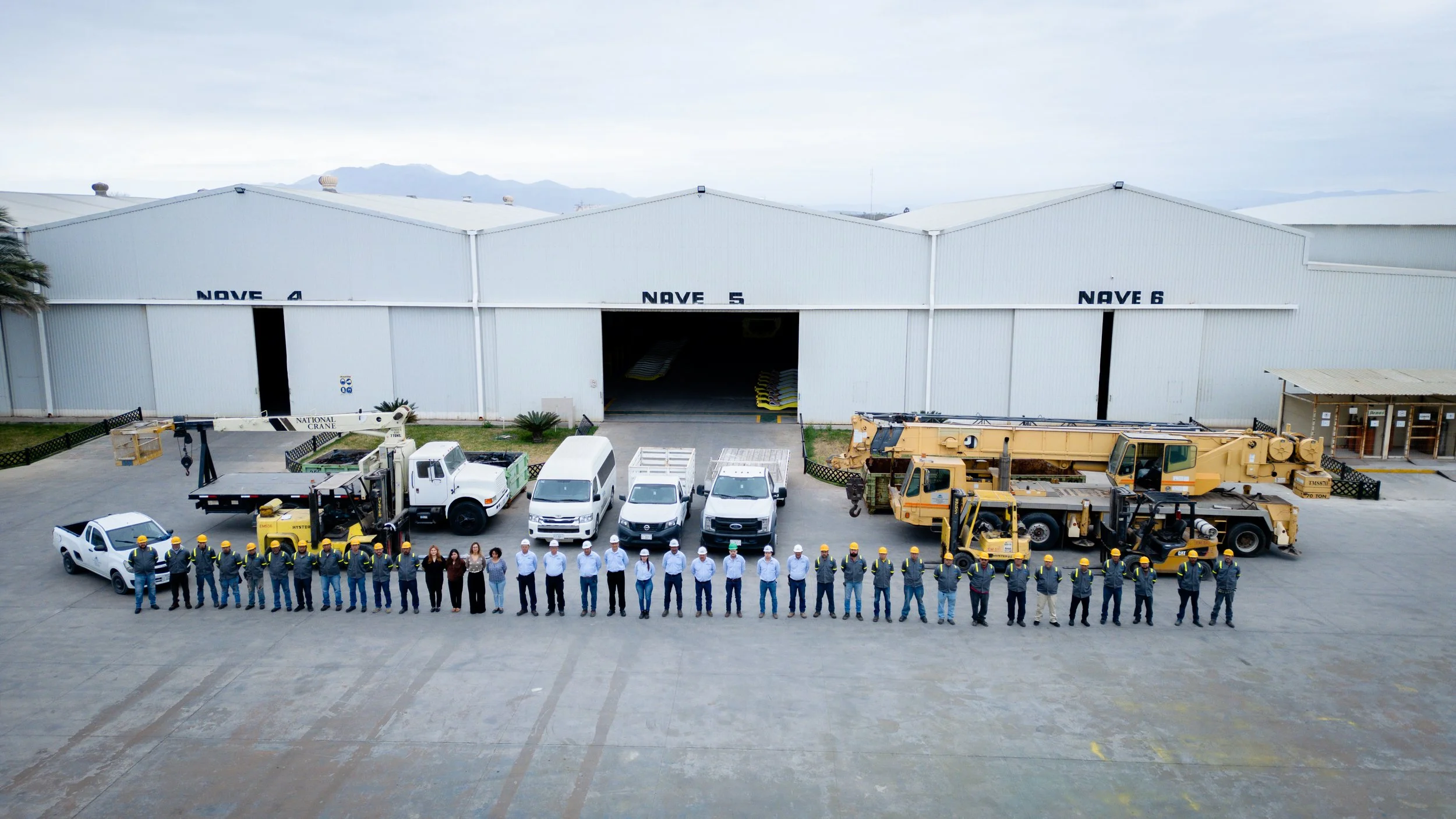 Grupo de personas y maquinaria en un hangar de aeropuerto, con puertas labeled NAVE 4, 5 y 6, con montacargas, camiones y vehículos, frente a montañas y cielo nublado.