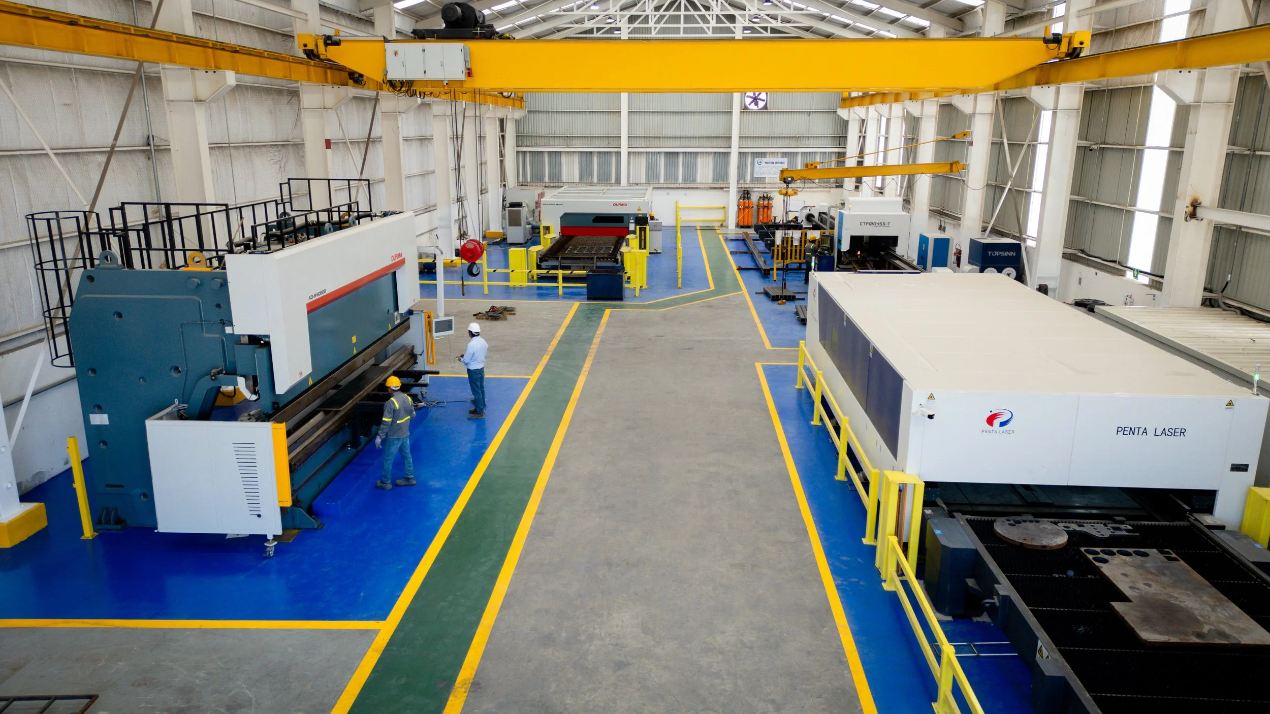 Interior de una fábrica con maquinaria industrial para procesamiento de láser, operarios con casco y uniforme de seguridad, estructura metálica y piso pintado en áreas delimitadas en azul, verde y amarillo.